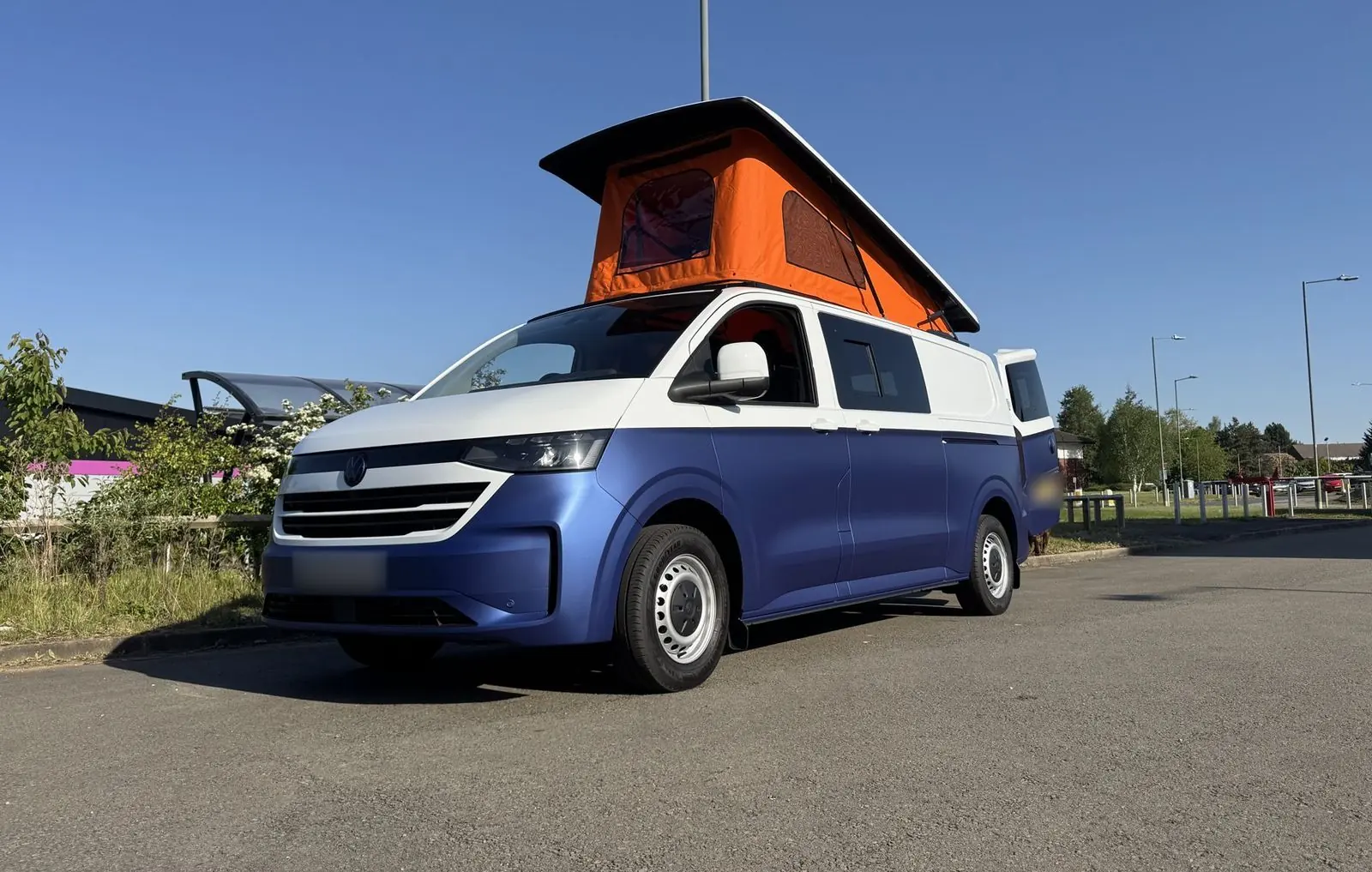 Front three-quarter view of a Volkswagen Transporter campervan in matt blue with a white upper half, parked on tarmac with its orange pop-top roof raised against a clear blue sky.