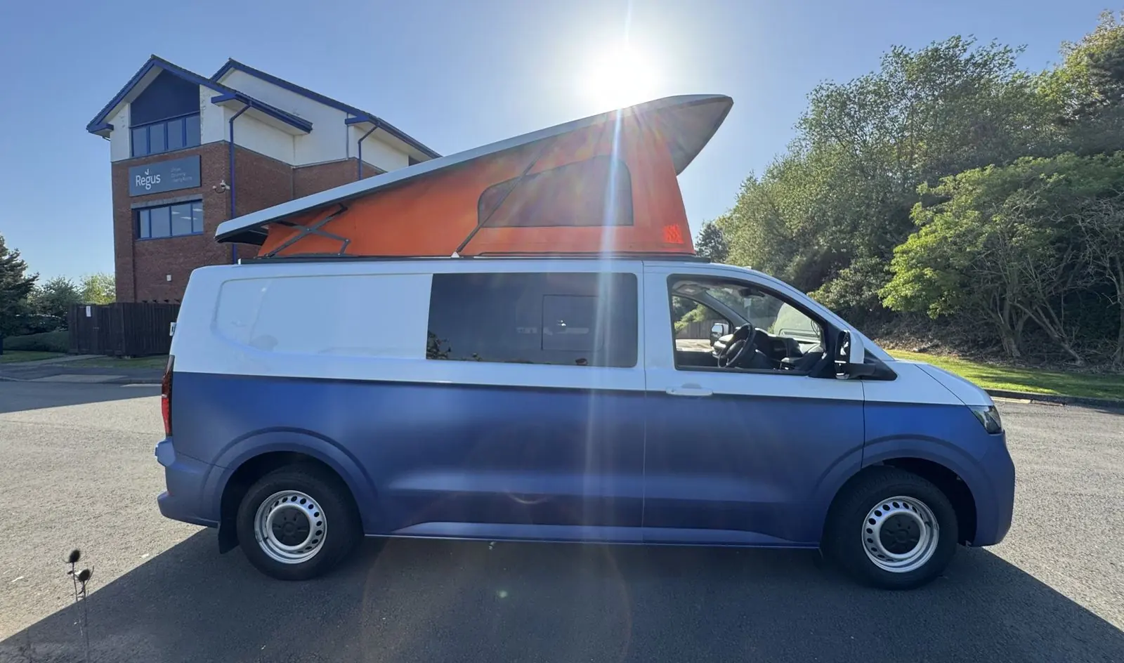 Side profile of the blue and white two-tone campervan with the orange pop-top raised, silver steel wheels and a tinted side window, with the sun directly behind the roof.