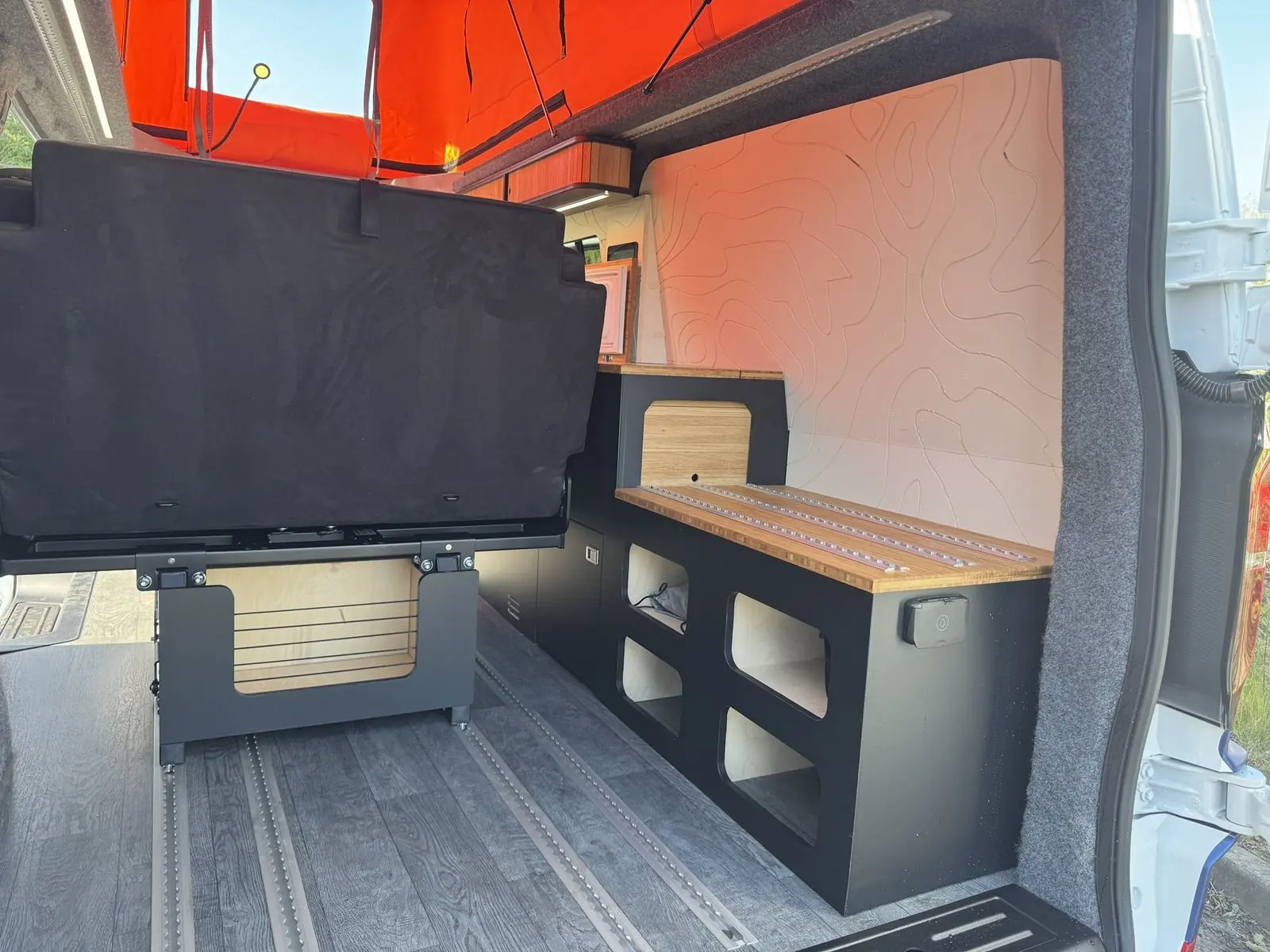 View in through the rear doors showing a black storage unit with open cubby holes and a bamboo top fitted with aluminium rails, a white contour-line wall panel, grey wood-effect floor with floor rails, and the back of the black rear seat.