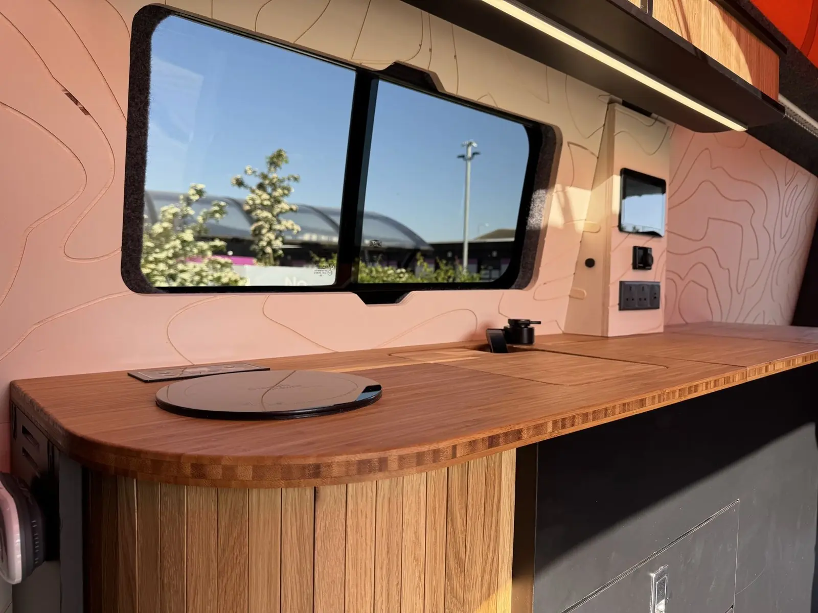 Bamboo kitchen worktop with a rounded end over slatted oak, a round black induction hob, a black tap over a covered sink, a sliding side window, and a touchscreen, dial controller and sockets on a contour-line wall panel that looks peach in the light coming through the orange roof canvas.