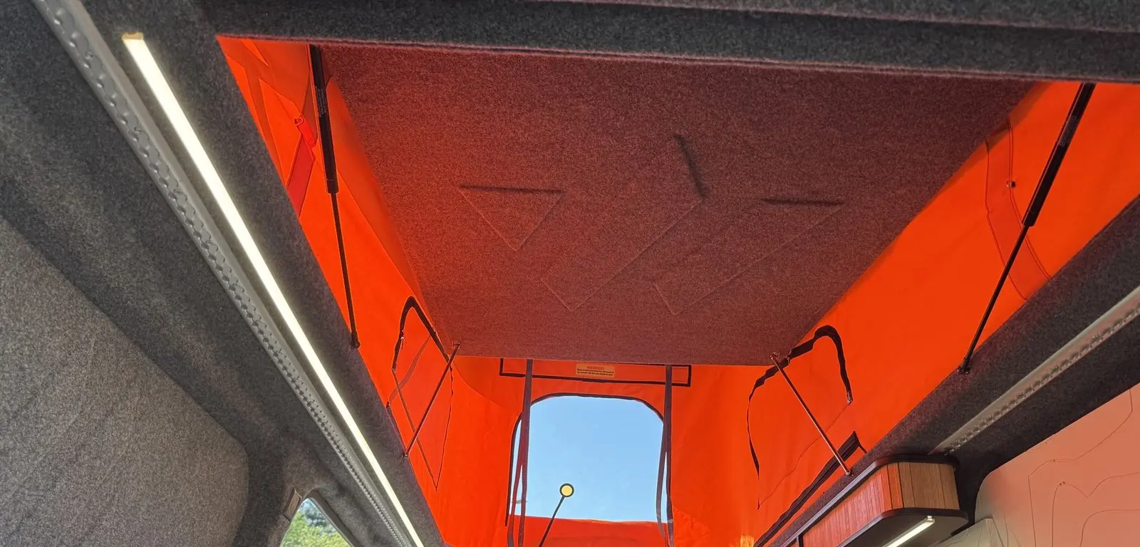 Looking up into the raised pop-top roof from inside the van, showing the orange canvas with a clear front window, grey carpet-lined roof and an LED strip light along the side.