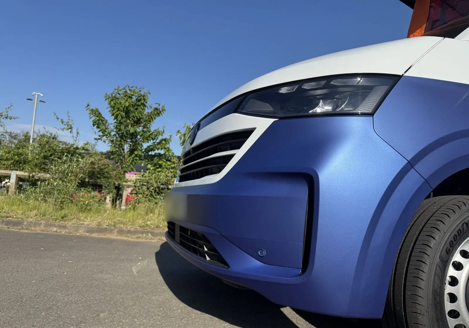 Low close-up of the front corner of the van showing the matt blue lower bodywork, white bonnet, LED headlight and silver steel wheel.