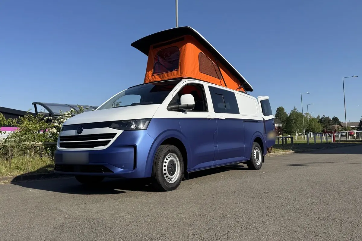 Front three-quarter view of a Volkswagen Transporter campervan in matt blue with a white upper half, parked on tarmac with its orange pop-top roof raised against a clear blue sky.