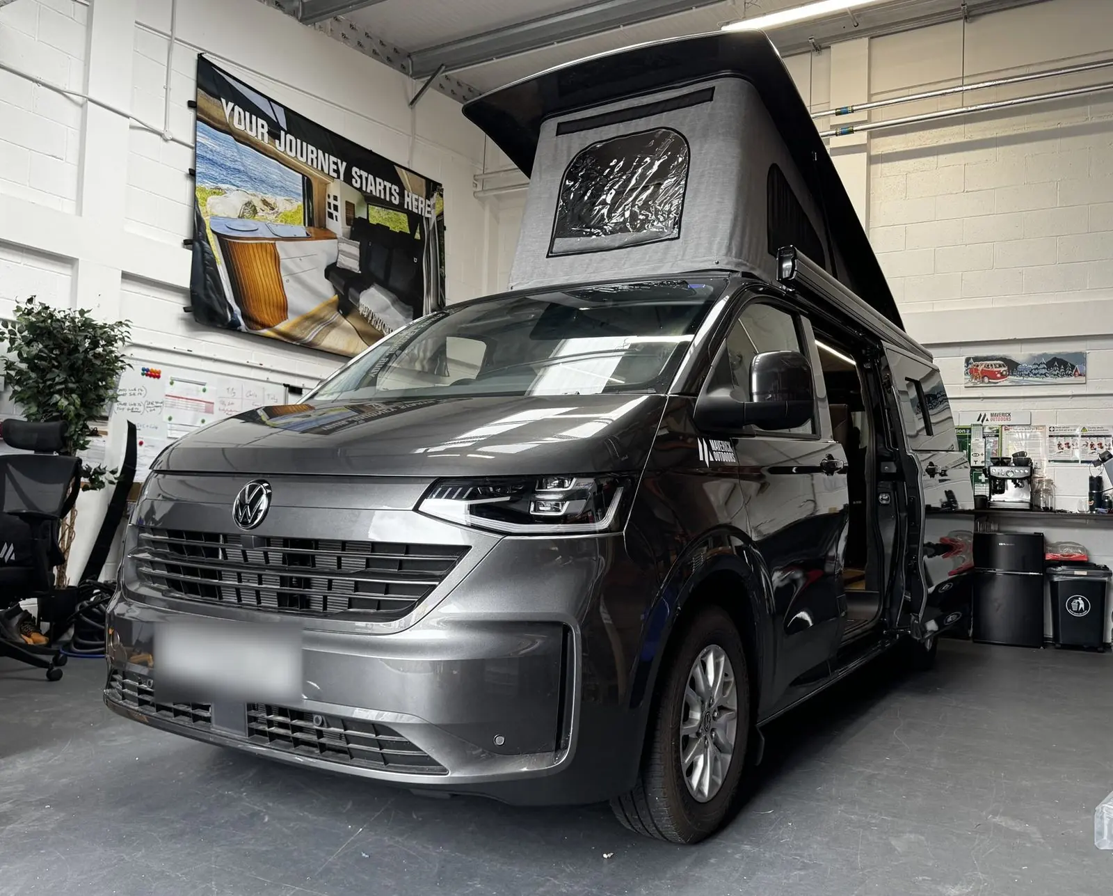 Front three-quarter view of a dark grey Volkswagen Transporter campervan inside the workshop with its pop-top roof raised, showing the black roof shell and light grey canvas.