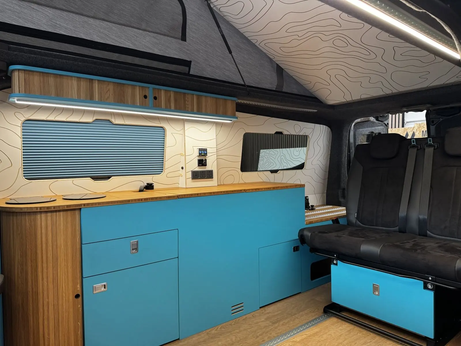 Campervan interior with a bright blue kitchen unit and bamboo worktop, slatted oak curved end panel, oak overhead locker with LED strip, blue pleated window blind, cream contour-line wall panels, a black rear seat and a contour-line ceiling panel.