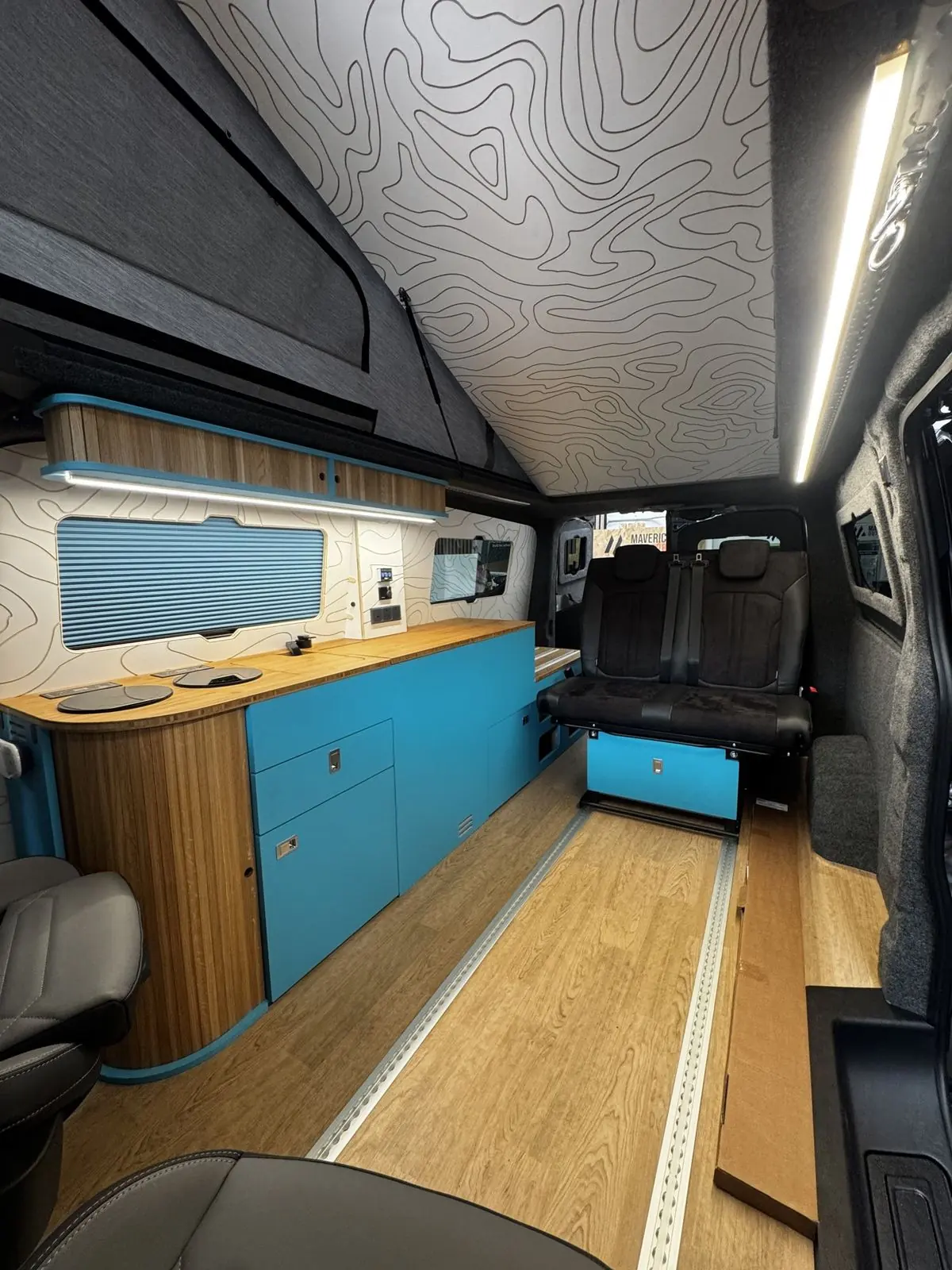 Tall view down the length of the interior from the front seats, showing the blue kitchen along one side, the black rear seat on a blue base, oak-effect floor with aluminium rails, and the white contour-line ceiling under the raised roof.