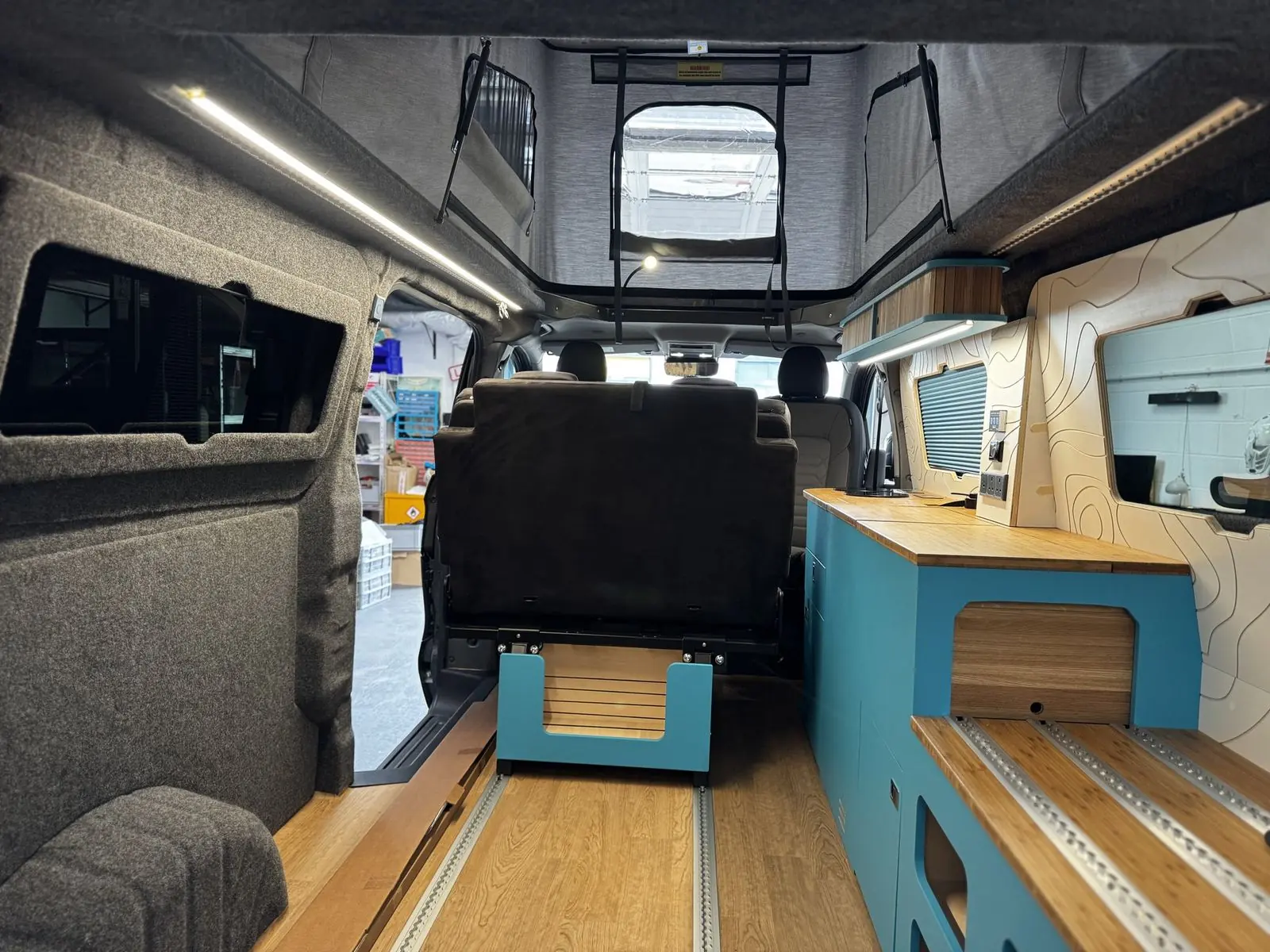 View from the rear of the van looking forward past a blue storage unit with a bamboo top and rails, the back of the rear seat on a blue frame, grey carpet-lined walls, and the raised pop-top with grey canvas.