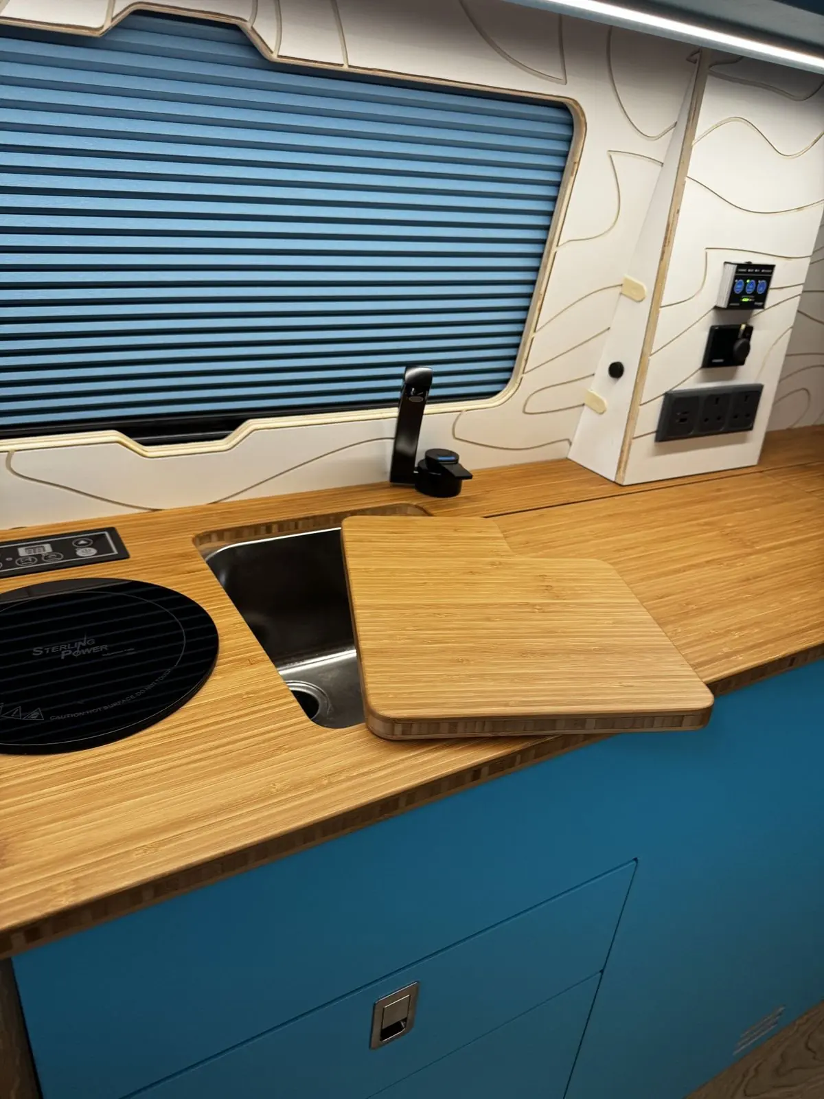 Stainless steel sink with a bamboo cover slid part-way off, black tap, blue pleated window blind, and a wall panel with a control panel, dial controller and sockets.