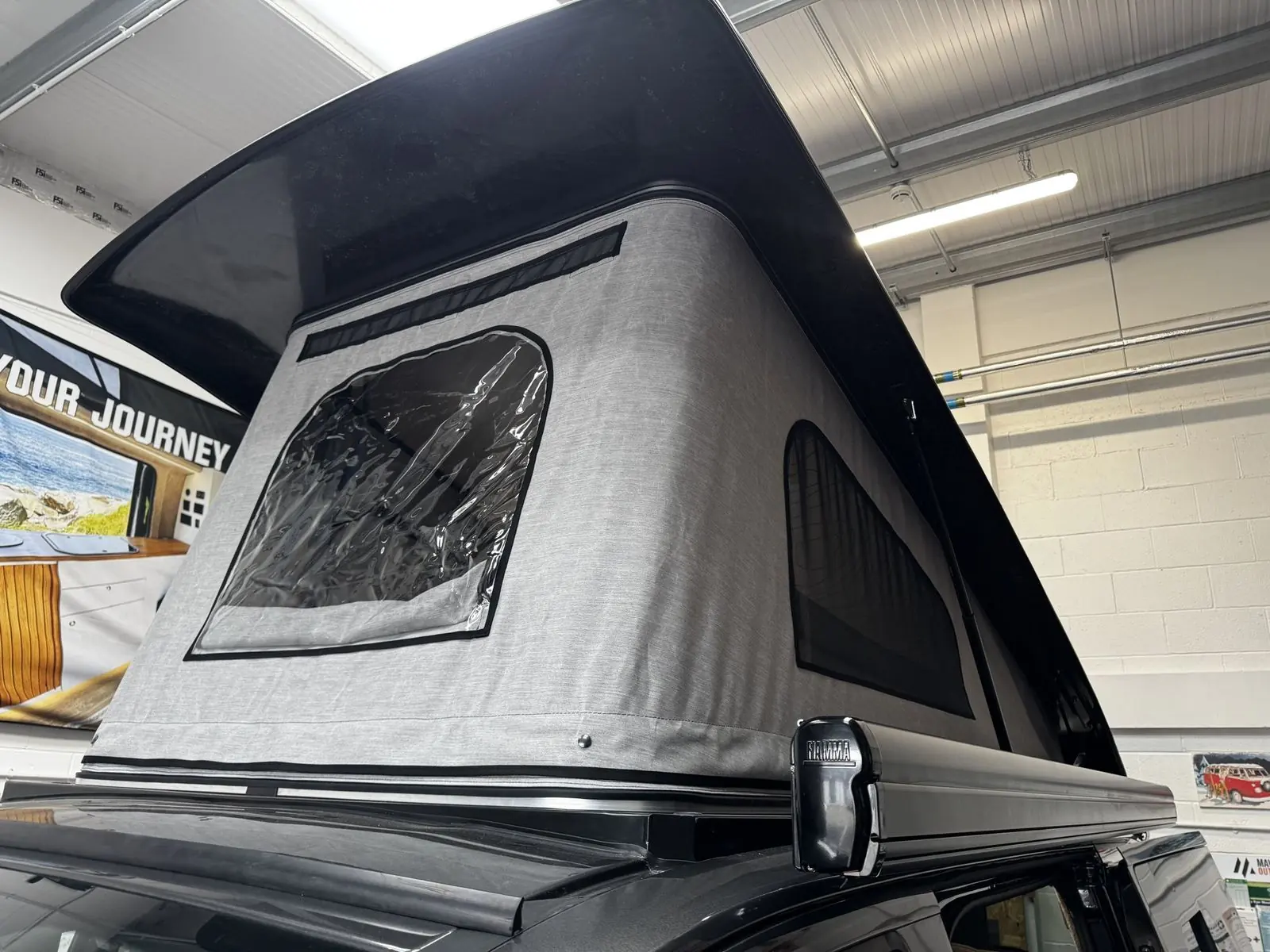 Close-up of the raised pop-top from outside, showing the gloss black roof shell, light grey canvas with a clear window and mesh vent, and a black Fiamma awning mounted along the roof edge.