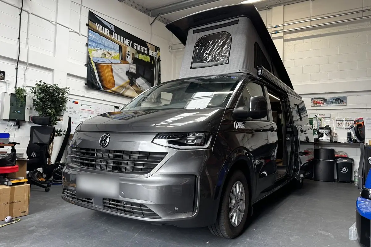 Front three-quarter view of a dark grey Volkswagen Transporter campervan inside the workshop with its pop-top roof raised, showing the black roof shell and light grey canvas.