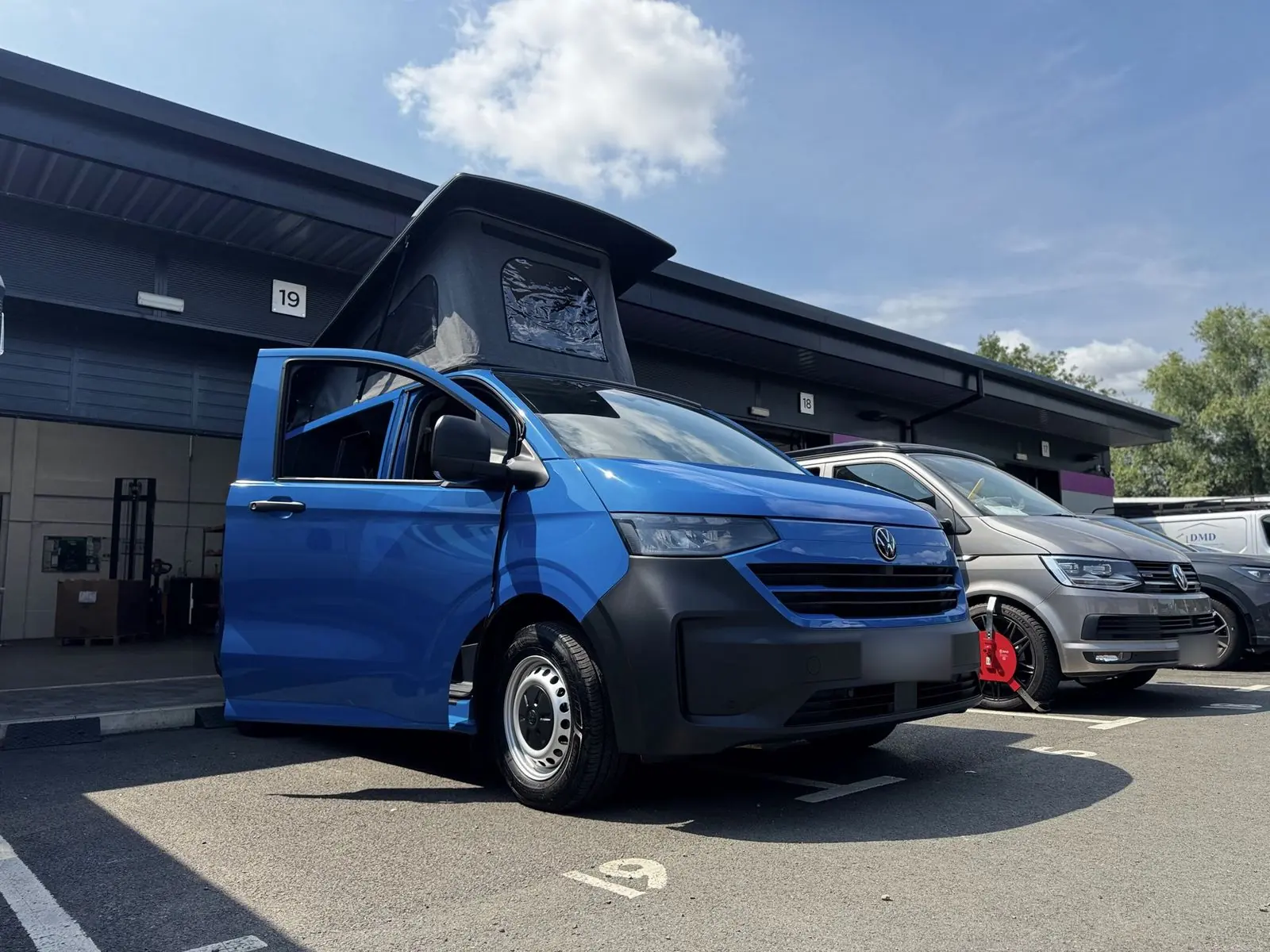Low front three-quarter view of a bright blue new-shape Volkswagen Transporter campervan with a dark grey lower bumper, silver steel wheels, the driver's door open and a dark grey pop-top roof raised.