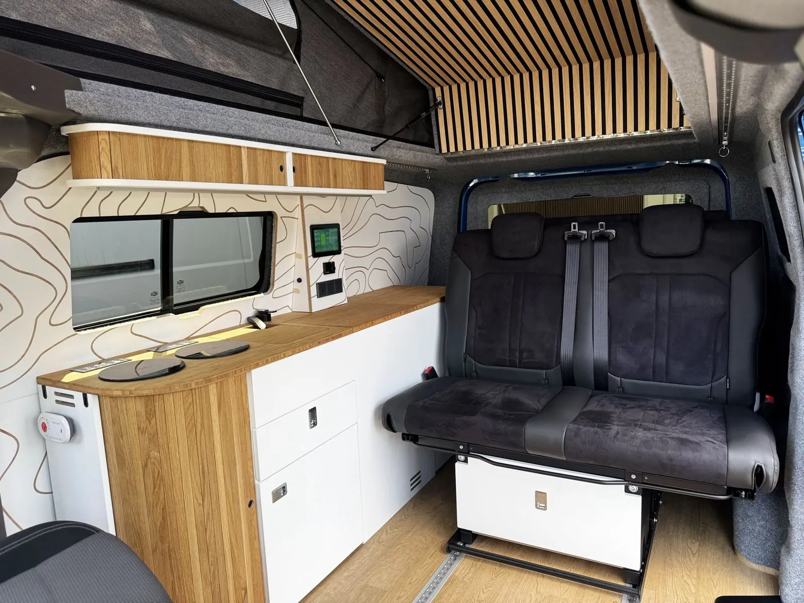 Interior with a white kitchen unit with an oak slatted curved end and bamboo worktop, a two-seat rear bench in black suede-effect fabric on a white base, an oak and white overhead locker, contour-line wall panels and a slatted roof panel.