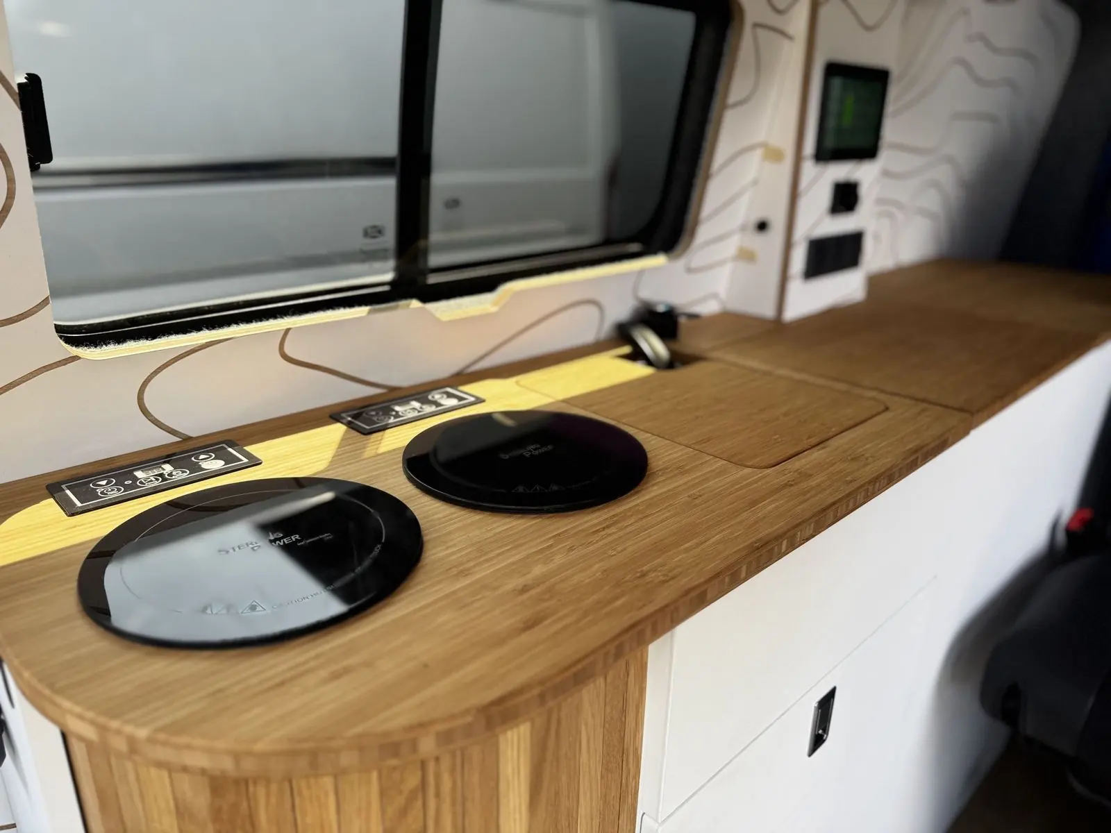 Bamboo worktop with two round black induction hobs with touch control panels, a flush bamboo sink cover and a black tap, with the touchscreen wall panel behind.