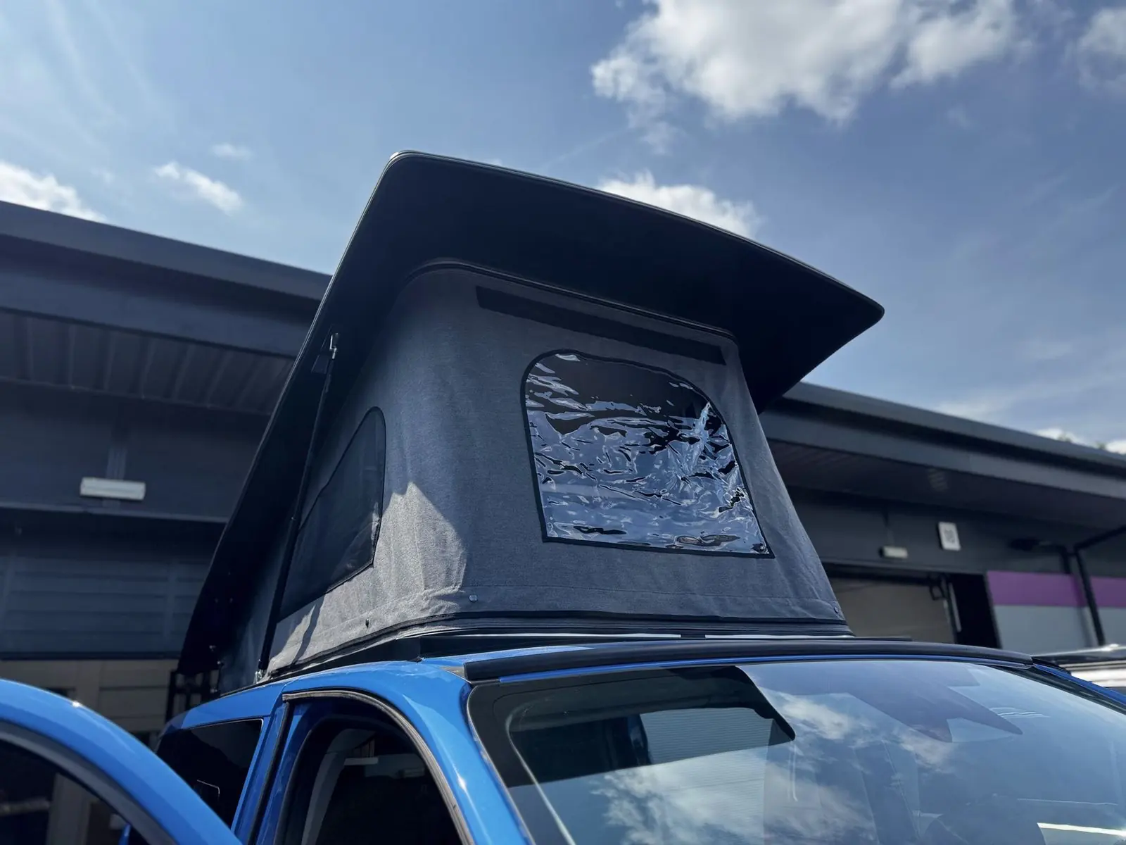 Raised pop-top roof with dark grey canvas, a clear window panel and a mesh side vent, above the blue cab roof.