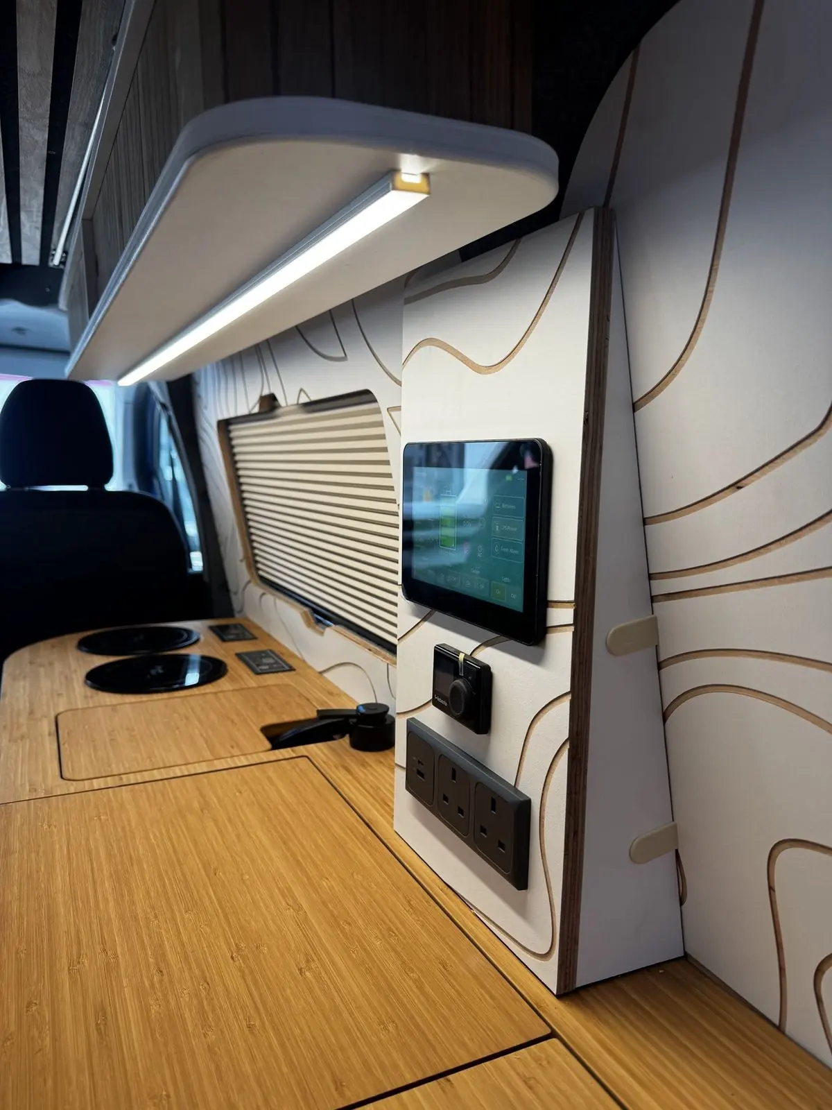 Looking along the bamboo worktop to a wall panel with a touchscreen, dial controller and double socket, a pleated window blind, two hobs and a white overhead locker with an LED strip light.