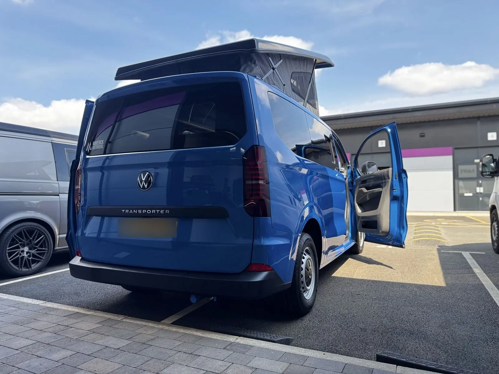 Rear three-quarter view of the bright blue campervan with a tailgate, tinted rear window, the pop-top raised and the passenger door open.
