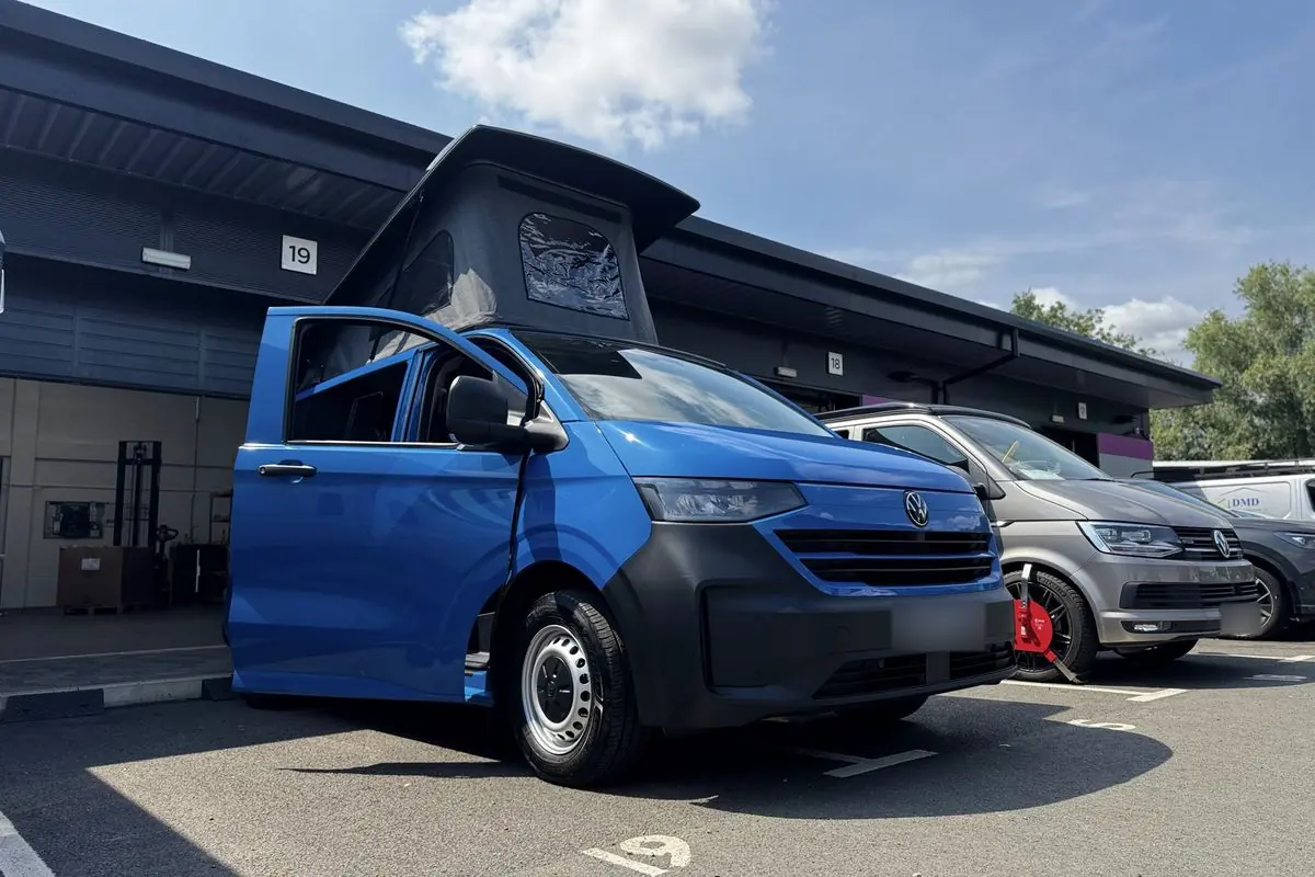 Low front three-quarter view of a bright blue new-shape Volkswagen Transporter campervan with a dark grey lower bumper, silver steel wheels, the driver's door open and a dark grey pop-top roof raised.