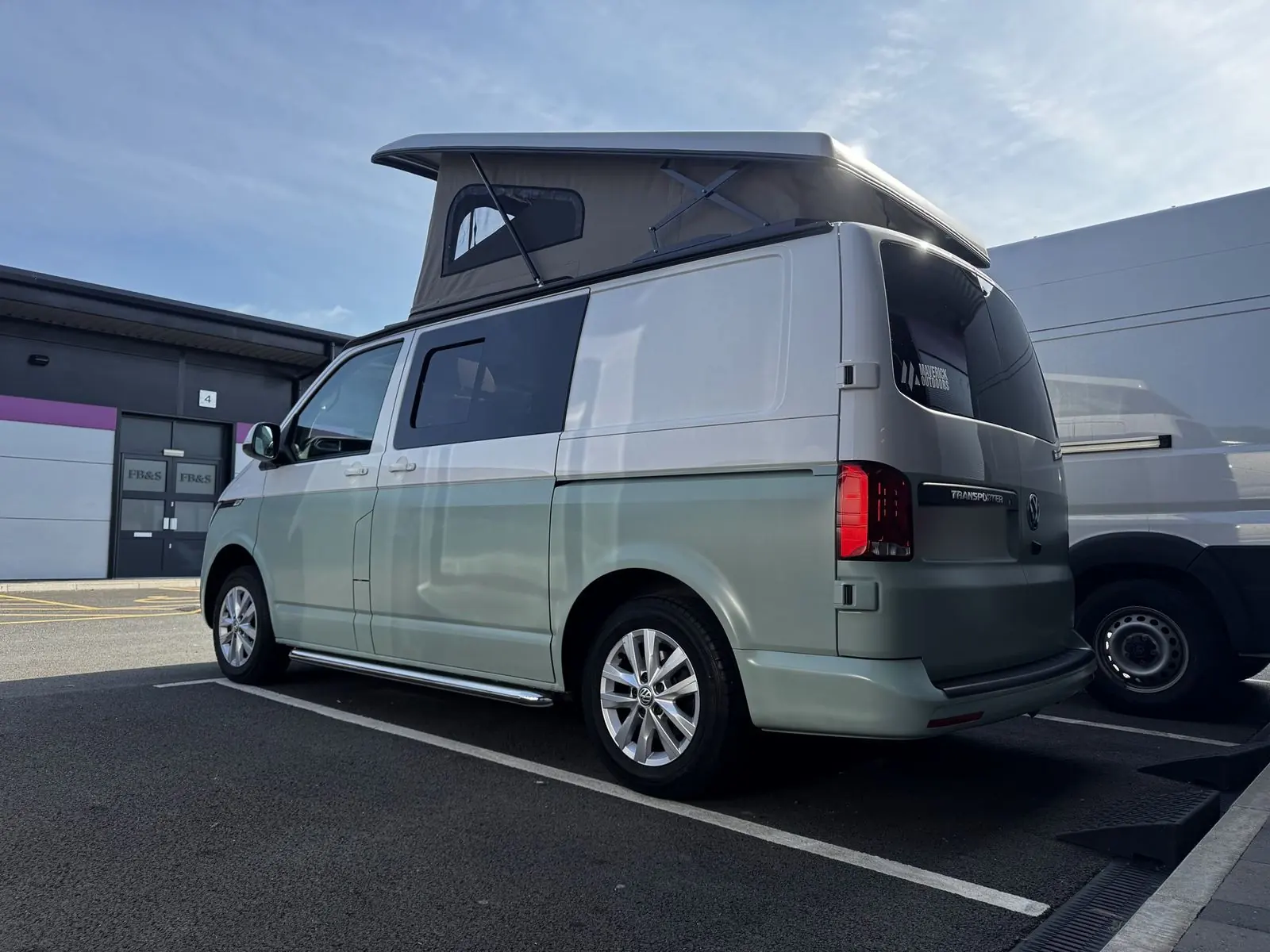 Rear three-quarter view of a Volkswagen Transporter campervan in two-tone paint, pale green lower half and light silver upper half, with a grey-beige pop-top roof raised, silver alloy wheels and chrome side bars.