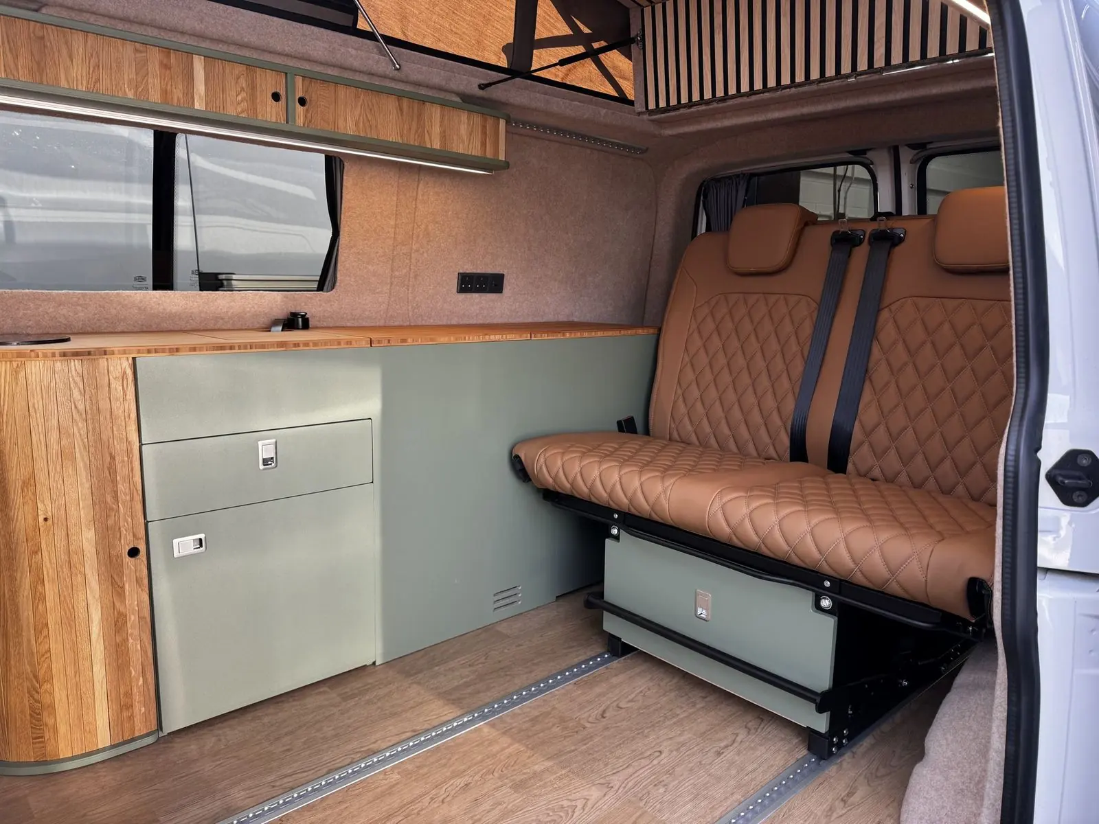 Interior with a tan diamond-quilted two-seat rear bench on a sage green base, a sage green kitchen unit with an oak slatted curved end and bamboo worktop, an oak overhead locker with LED strip, beige carpet-lined walls and a slatted roof panel.