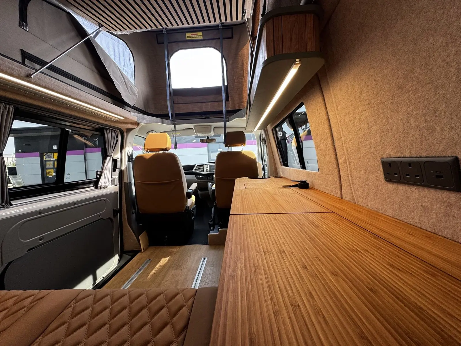View forward from the rear seat along the bamboo worktop to two tan front seats, with beige carpet-lined walls, a triple socket panel, an overhead locker with LED strip and the raised pop-top with a front window panel.