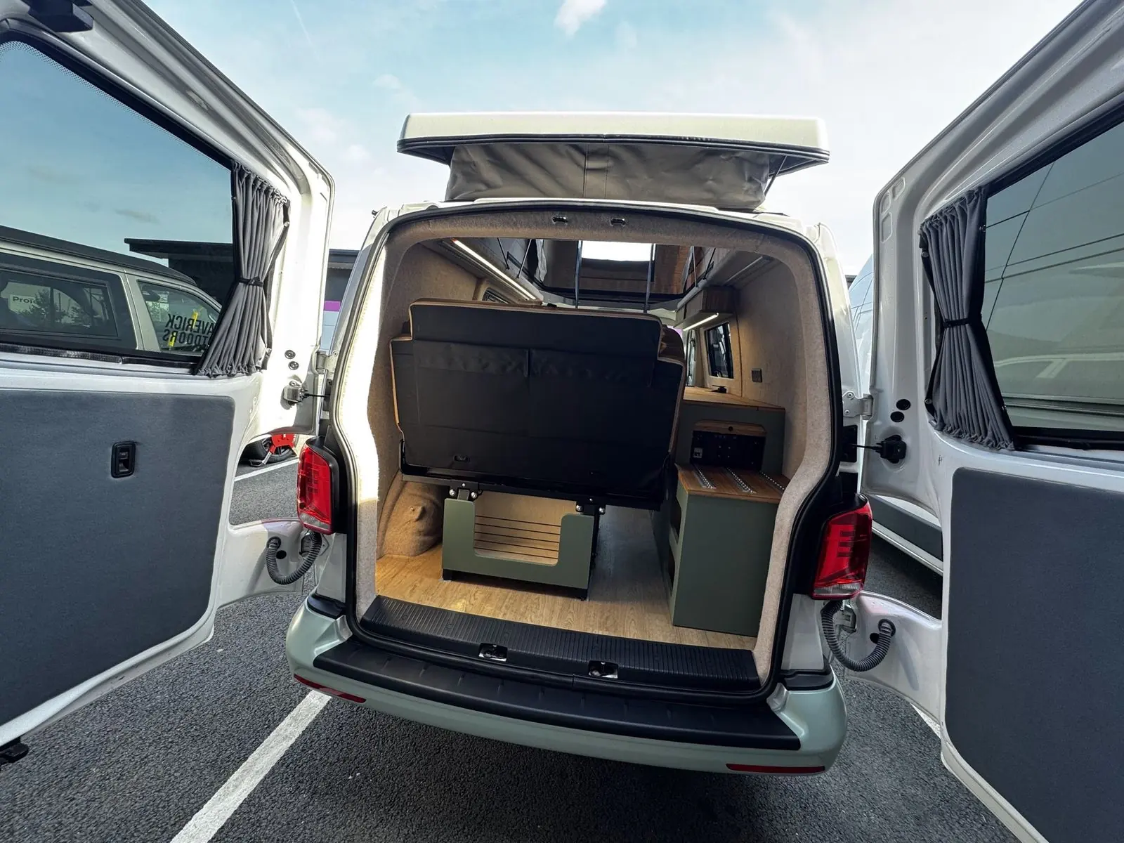 View in through the open rear barn doors showing the back of the rear seat, a sage green rear storage unit with a bamboo top and rails, grey curtains on the door windows and the pop-top raised.