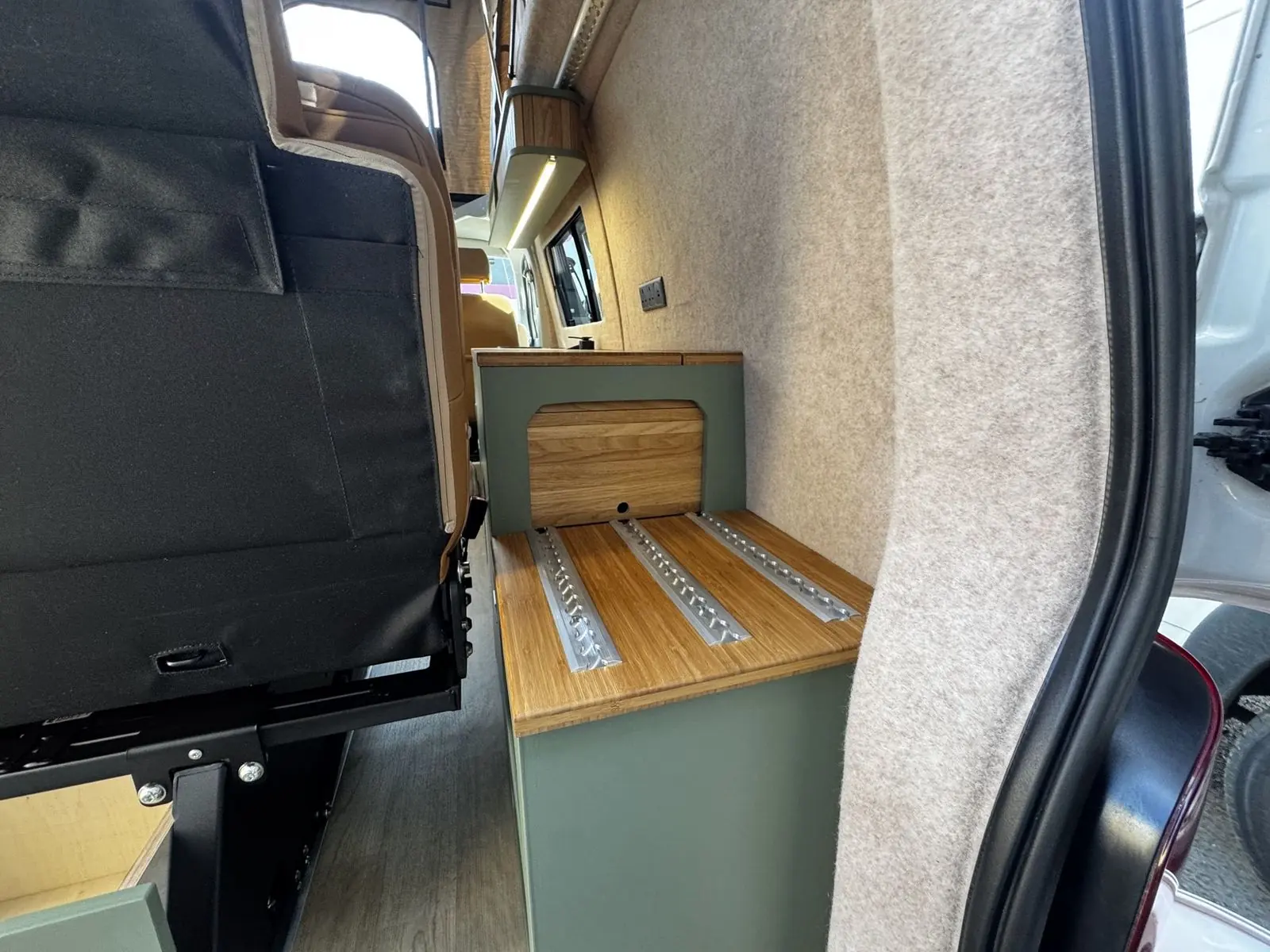 Rear storage unit in sage green with a bamboo top fitted with three aluminium rails and an oak tambour door, beside beige carpet-lined walls.