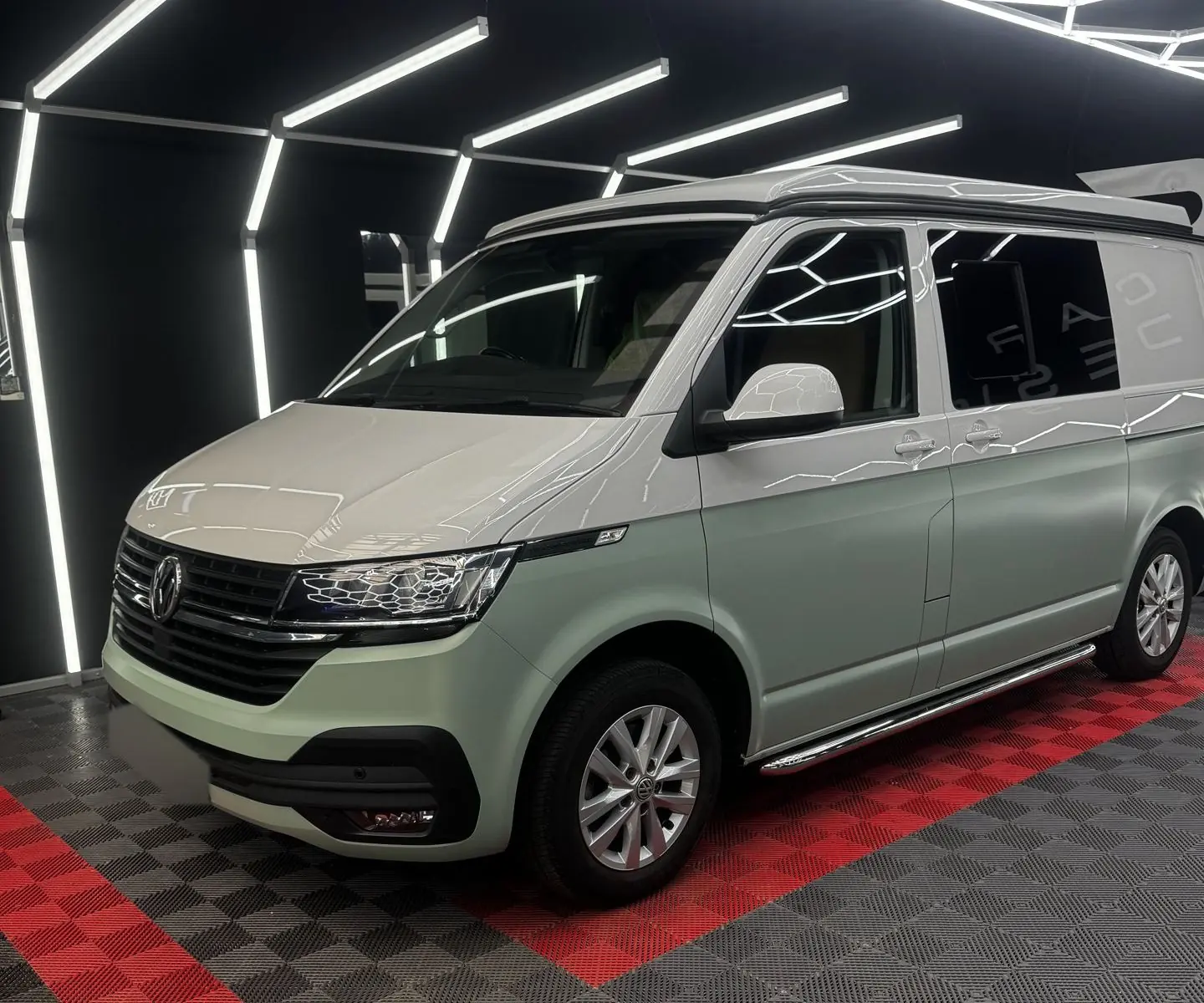 Front three-quarter view of the pale green and white-silver two-tone Volkswagen Transporter with the pop-top closed, photographed indoors under hexagonal strip lights on a black and red tiled floor.