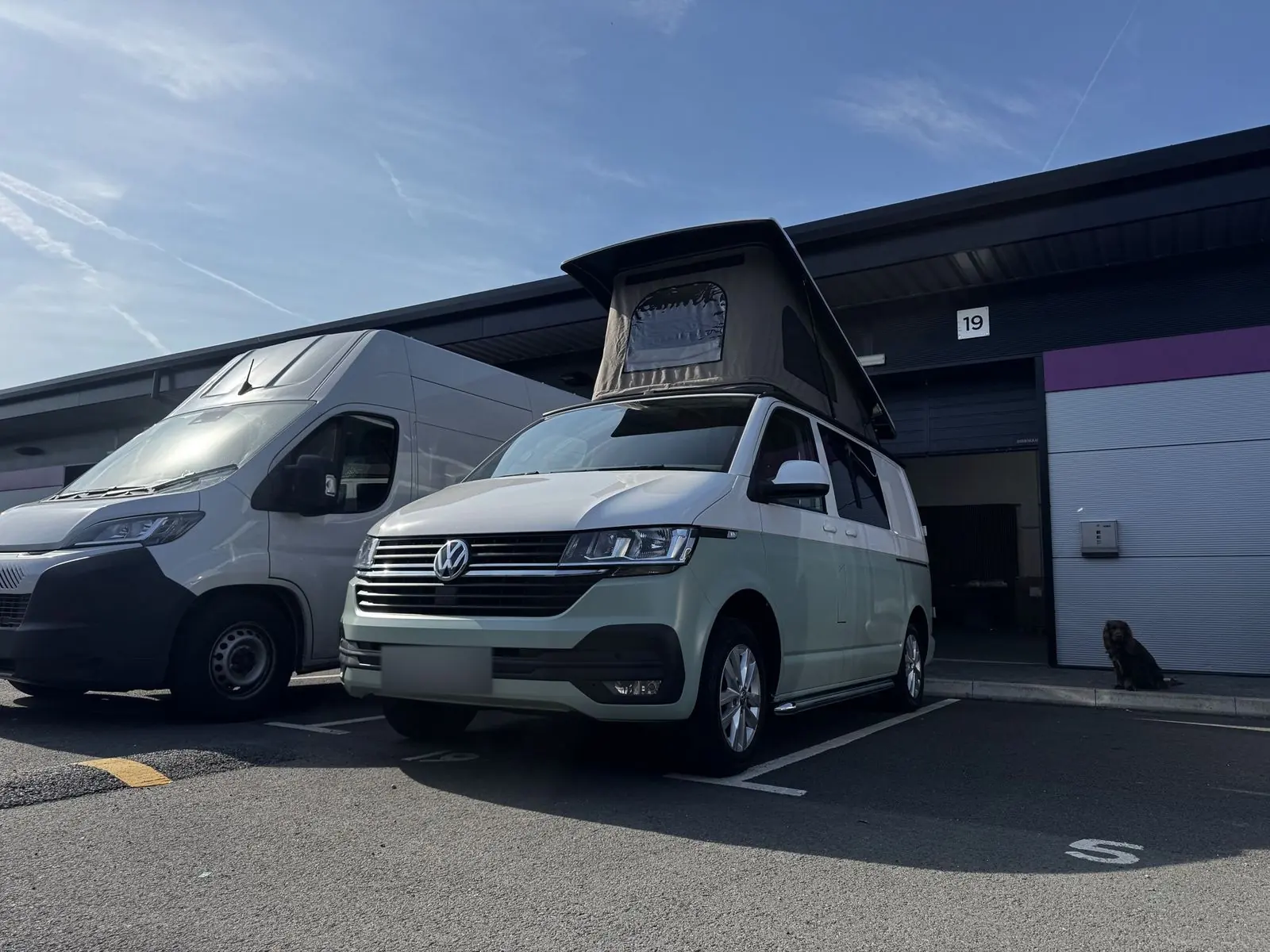Low front three-quarter view of the two-tone campervan parked outside a business unit with its grey-beige pop-top raised.