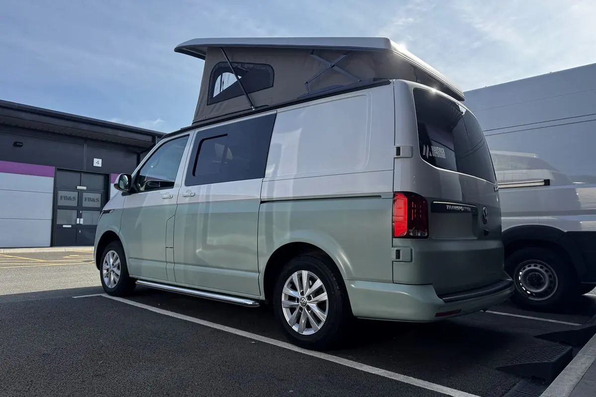 Rear three-quarter view of a Volkswagen Transporter campervan in two-tone paint, pale green lower half and light silver upper half, with a grey-beige pop-top roof raised, silver alloy wheels and chrome side bars.