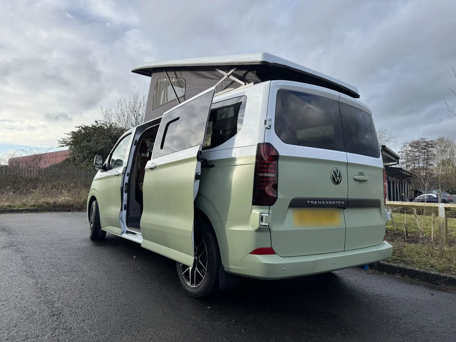 Rear three-quarter view of a Volkswagen Transporter campervan in matt light green with a white upper half, pop-top roof raised and side door slid open, parked on wet tarmac under a cloudy sky.