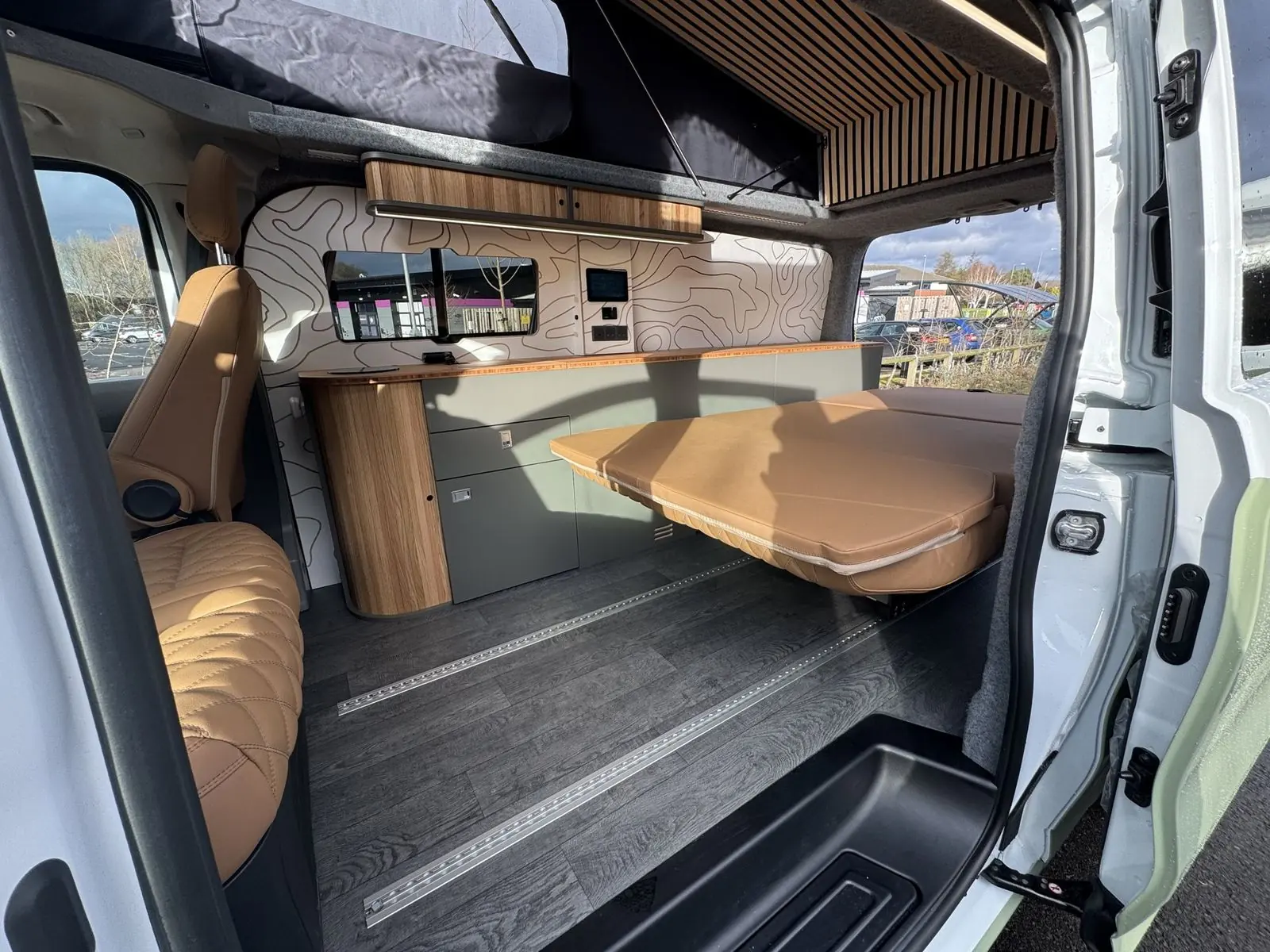View through the open side door of the interior with the rear seat folded flat into a tan bed, a sage green kitchen unit with bamboo worktop, white contour-line wall panels, tan quilted front seat and dark grey wood-effect floor with rails.