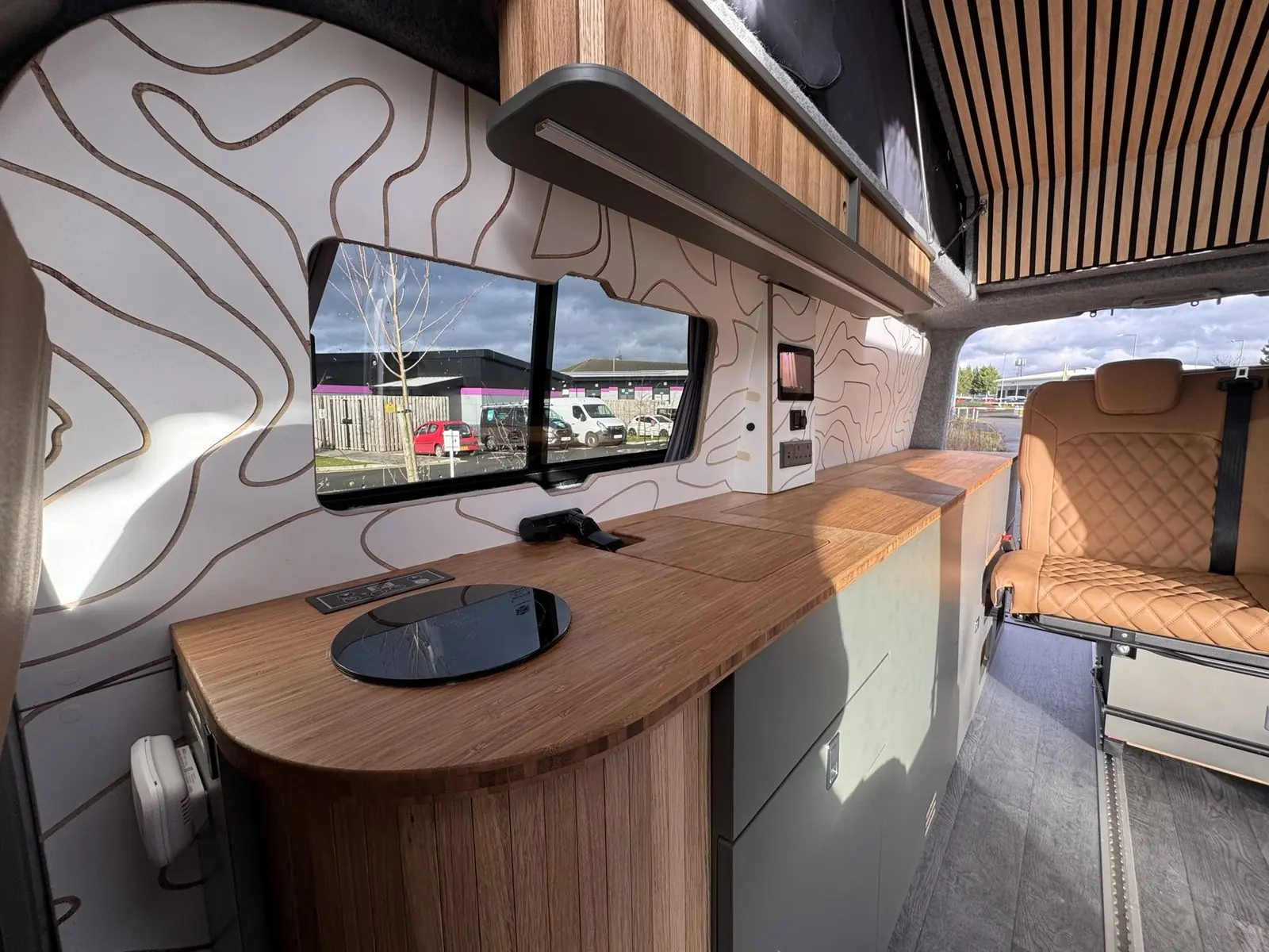 Bamboo kitchen worktop with a rounded end over slatted oak, a round black induction hob and black tap, below a sliding window and white contour-line wall panel, with the tan diamond-quilted rear seat beyond.
