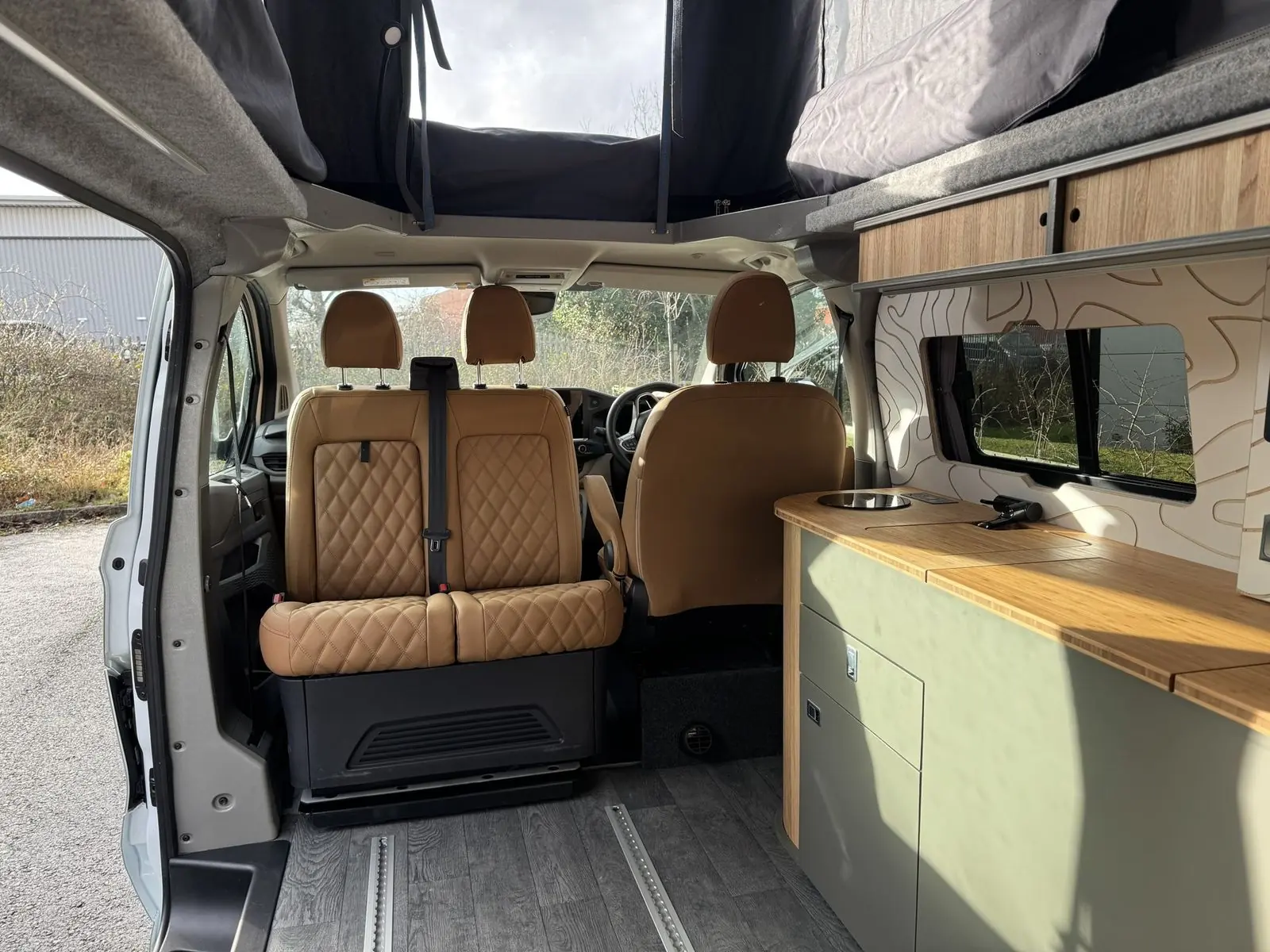 Looking forward inside the van at a tan diamond-quilted double front seat facing the living area, with the sage green kitchen unit on the right and the pop-top open above.