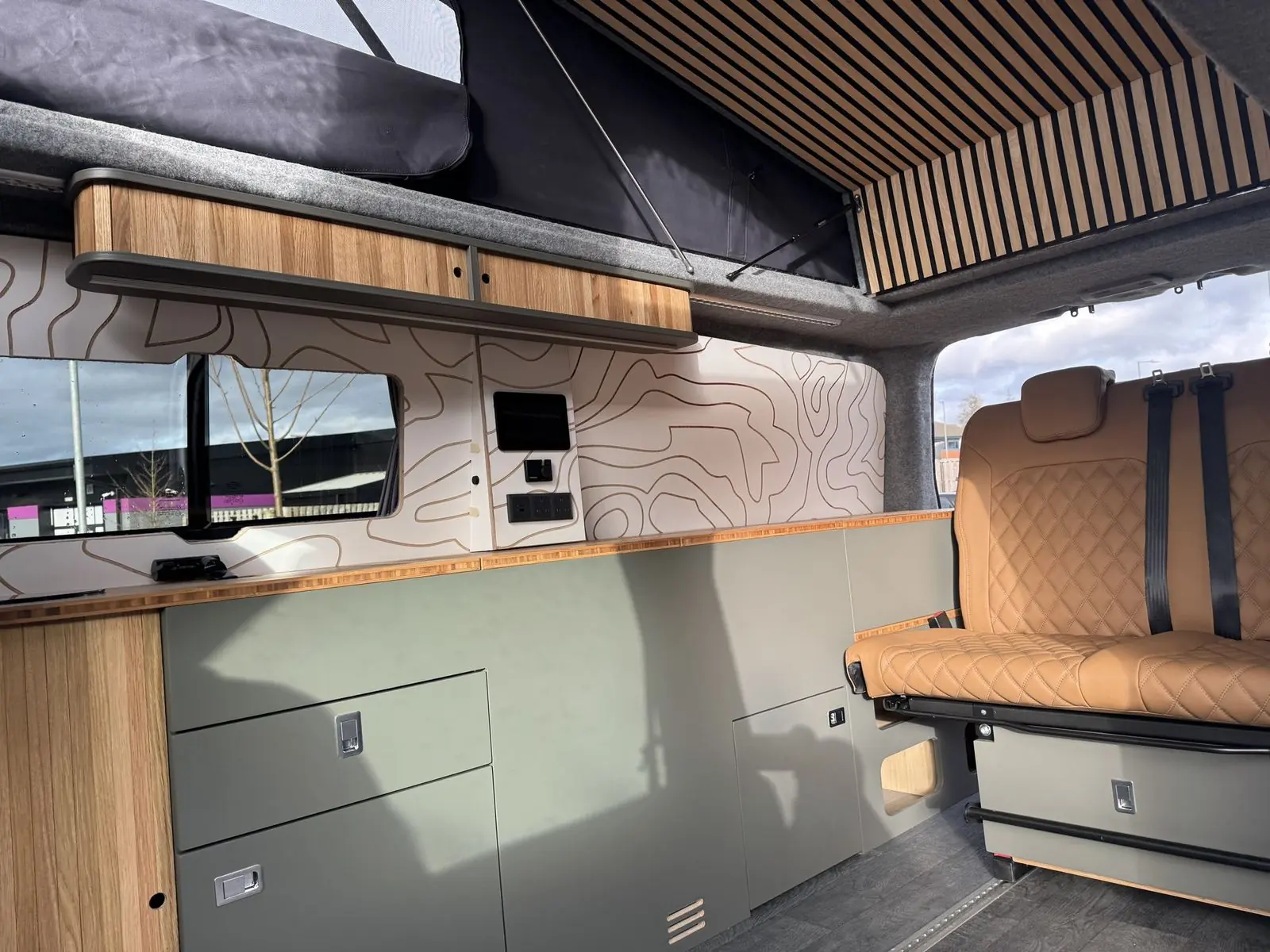Sage green kitchen cabinets with drawers under a bamboo worktop, oak overhead locker, wall-mounted touchscreen and sockets, tan quilted rear seat, and a slatted wood panel in the raised roof.