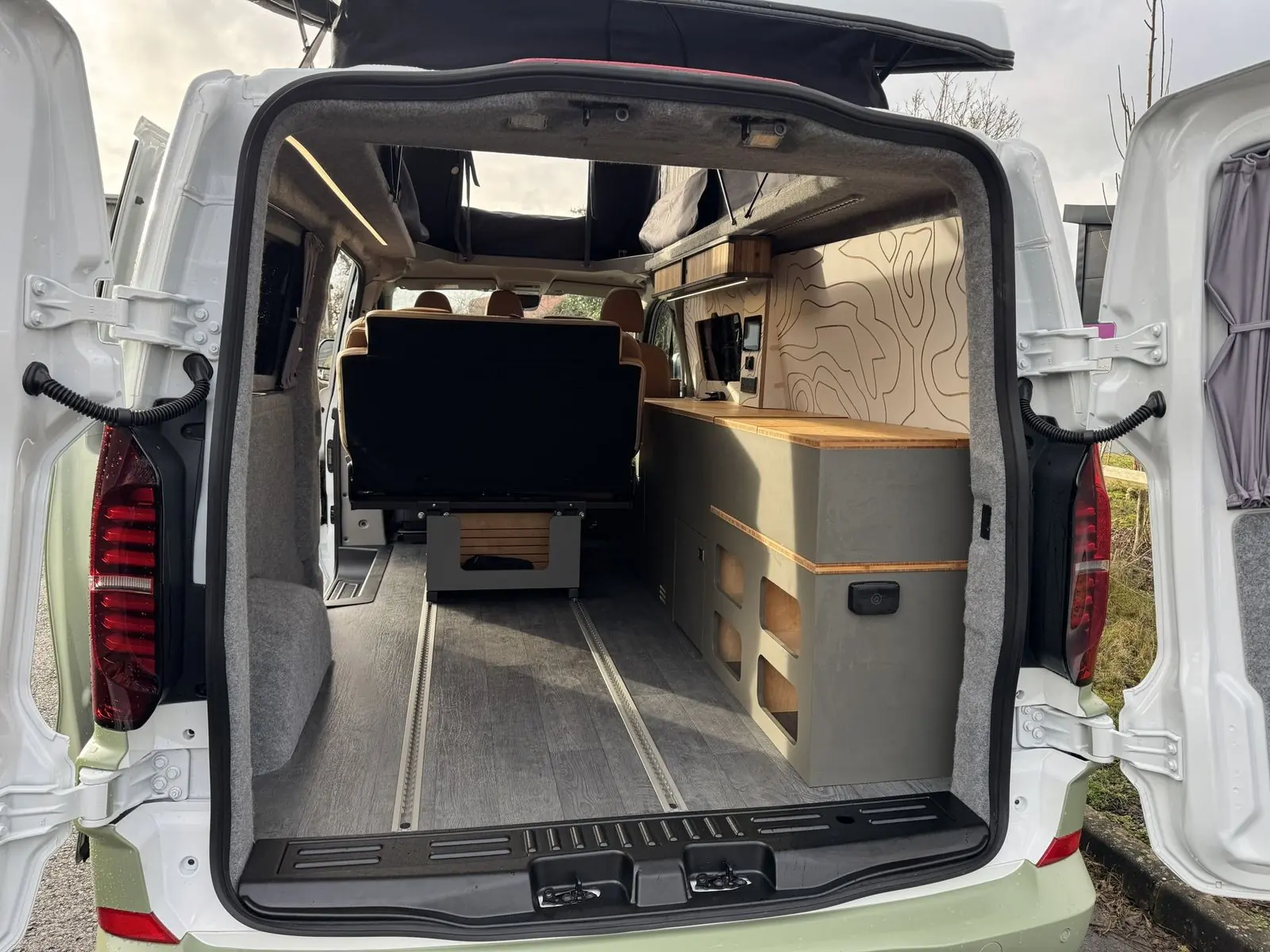 View in through the open rear doors showing the dark grey floor with two rails, the back of the rear seat, a sage green storage unit with open cubby holes and bamboo top, and grey carpet-lined walls.