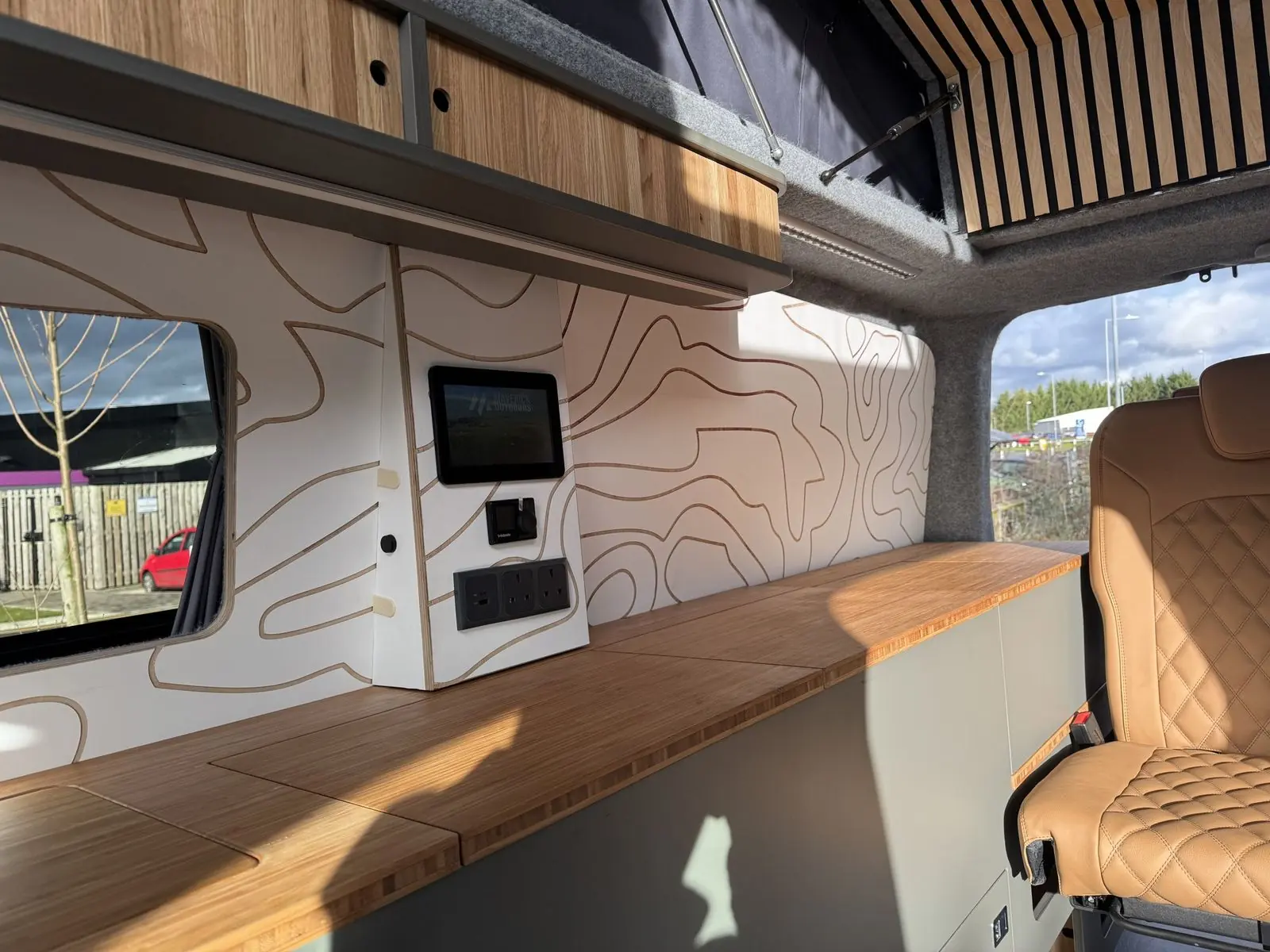 Close-up of the wall control panel with a touchscreen showing the Maverick Outdoors logo, a dial controller and a triple socket, above the bamboo worktop.