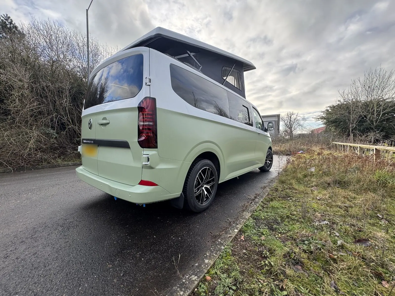 Rear view from the passenger side of the light green and white campervan with the pop-top raised, showing dark tinted windows and black and silver alloy wheels.
