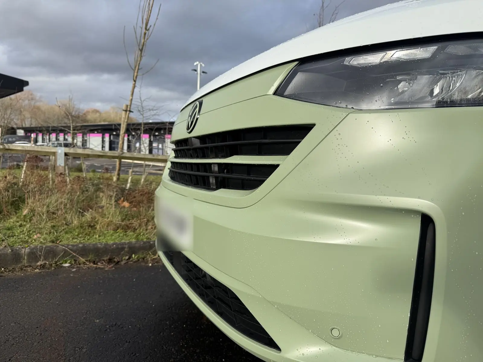 Low close-up of the front of the van showing the matt light green bumper and grille surround, LED headlight and white bonnet, with raindrops on the bodywork.