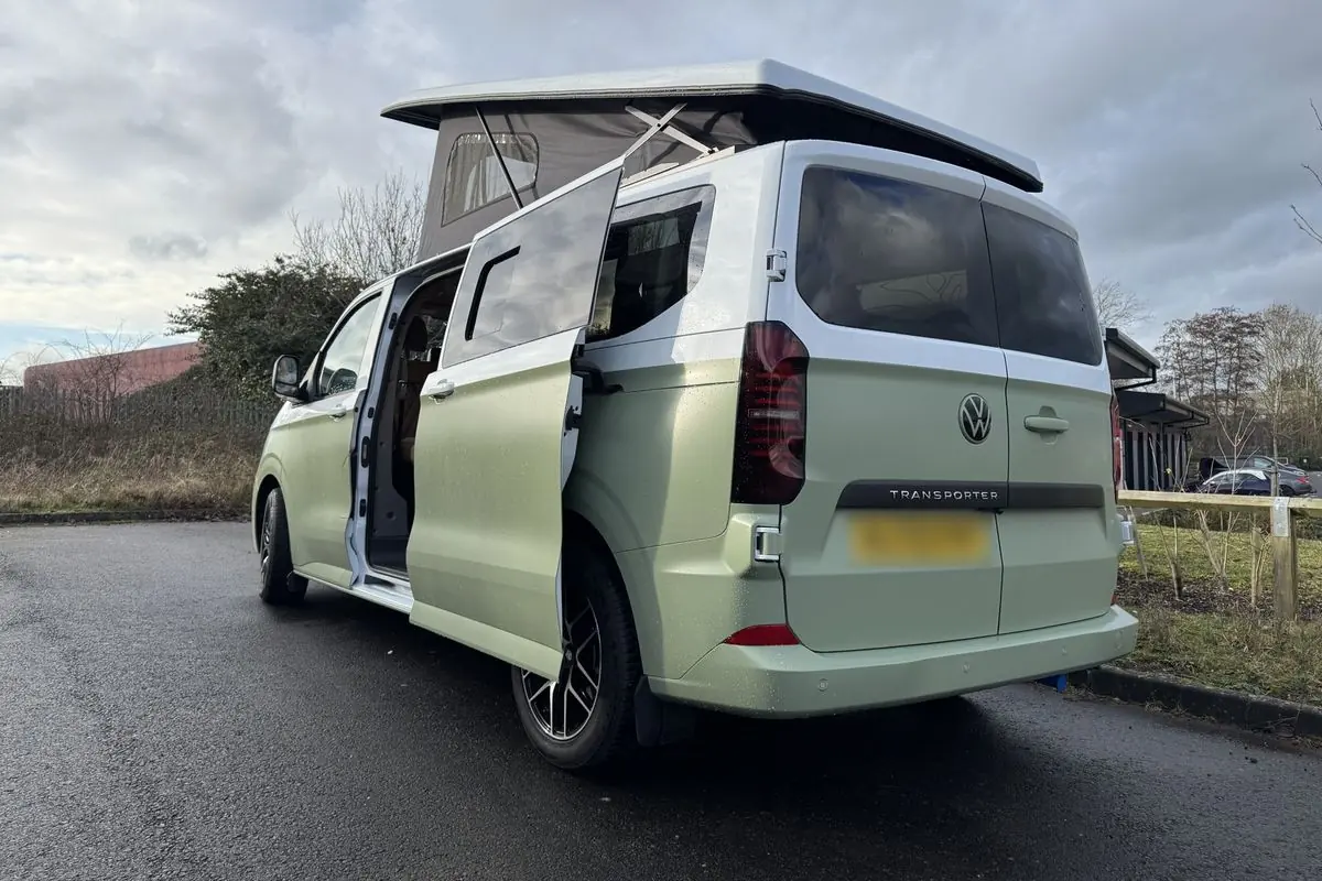 Rear three-quarter view of a Volkswagen Transporter campervan in matt light green with a white upper half, pop-top roof raised and side door slid open, parked on wet tarmac under a cloudy sky.