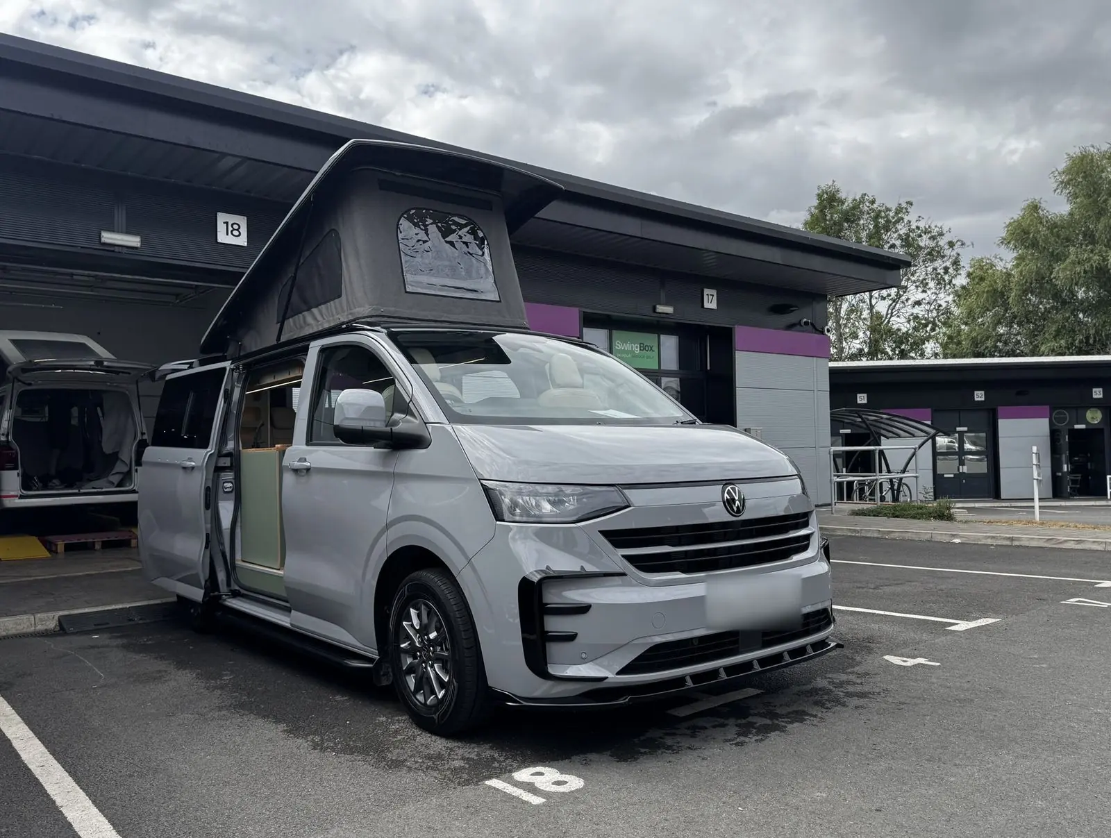 Front three-quarter view of a light grey new-shape Volkswagen Transporter campervan parked outside a workshop unit, with its dark grey pop-top roof raised, a side door slid open and a black front splitter.