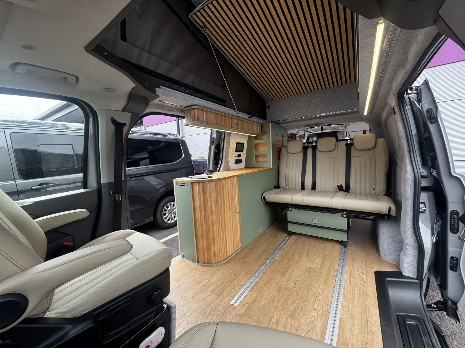 Wide interior view from the front seats showing a cream leather three-seat rear bench, a sage green kitchen pod with a slatted oak curved end and bamboo worktop, oak-effect floor with two rails, and a slatted wood panel in the raised roof.