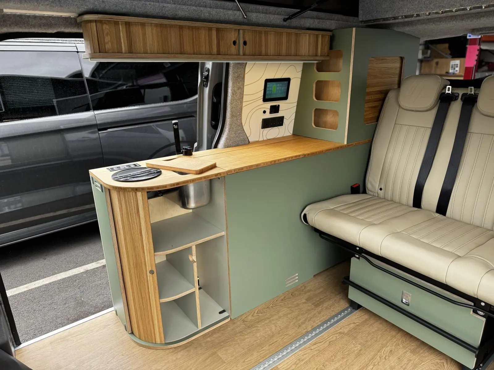 Sage green kitchen pod with its curved tambour door open to show shelves and a stainless steel sink bowl, a bamboo worktop with a round hob, an oak overhead locker, a touchscreen on a contour-line panel, and the cream leather rear bench.
