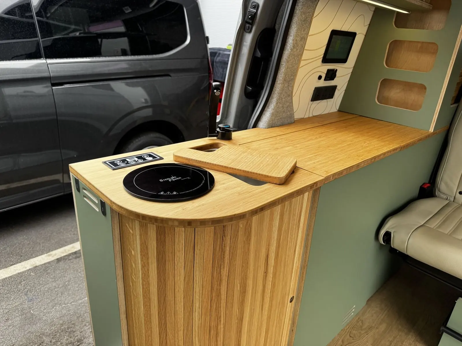 Close-up of the bamboo worktop with a round black Sterling Power induction hob and touch control panel, a bamboo sink cover, a black tap, and the slatted oak curved end of the kitchen pod.