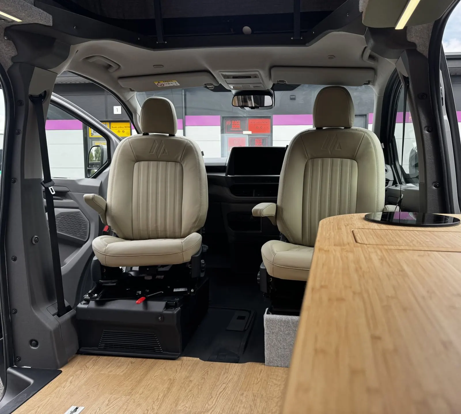 Two cream leather front seats with vertical stitched panels, armrests and an embossed logo, seen from the living area across the bamboo worktop.