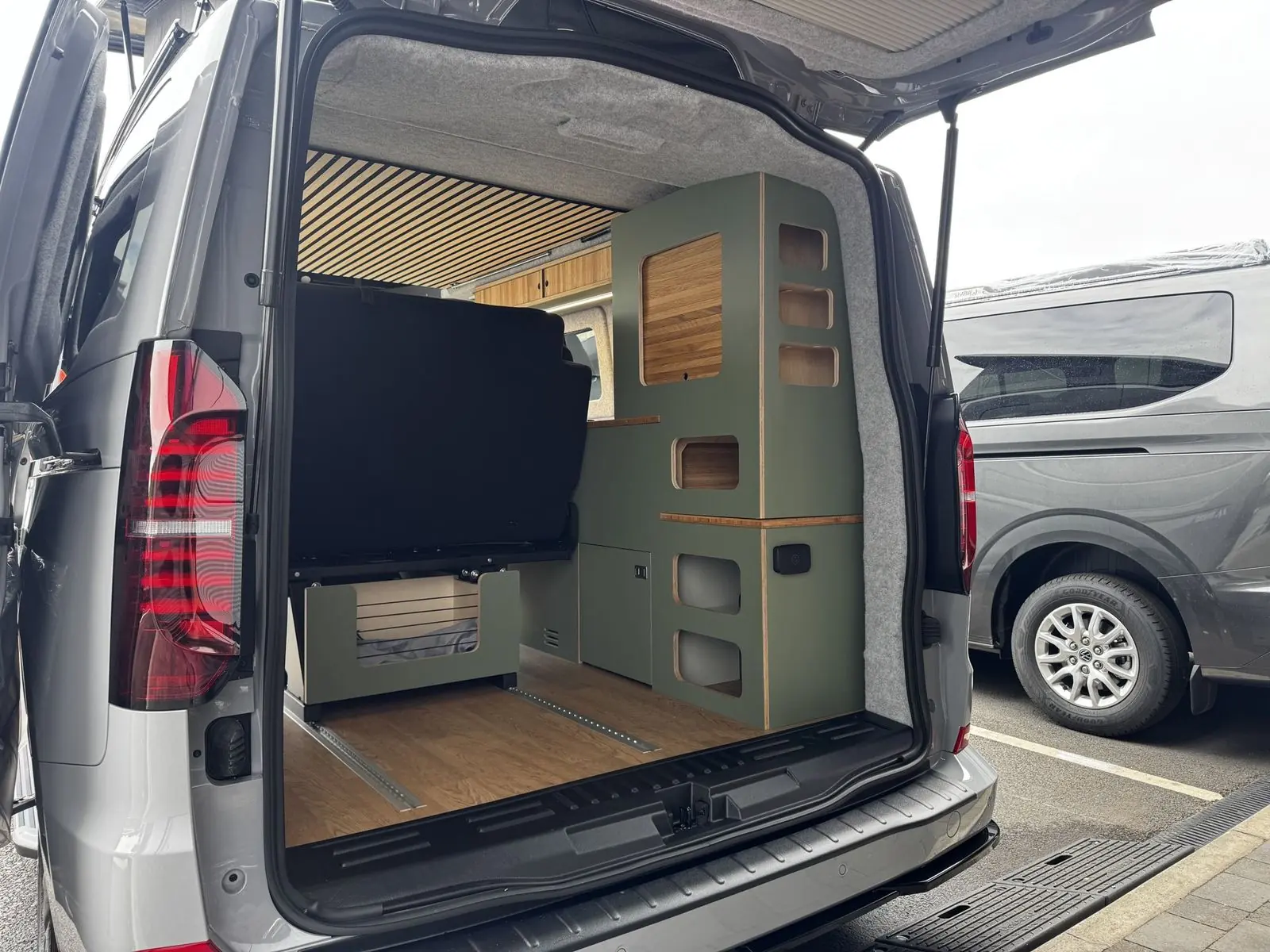 View in through the open tailgate showing a tall sage green storage unit with rounded cut-out cubby holes and an oak tambour door, the back of the rear seat, oak-effect floor with rails, and grey carpet-lined walls.