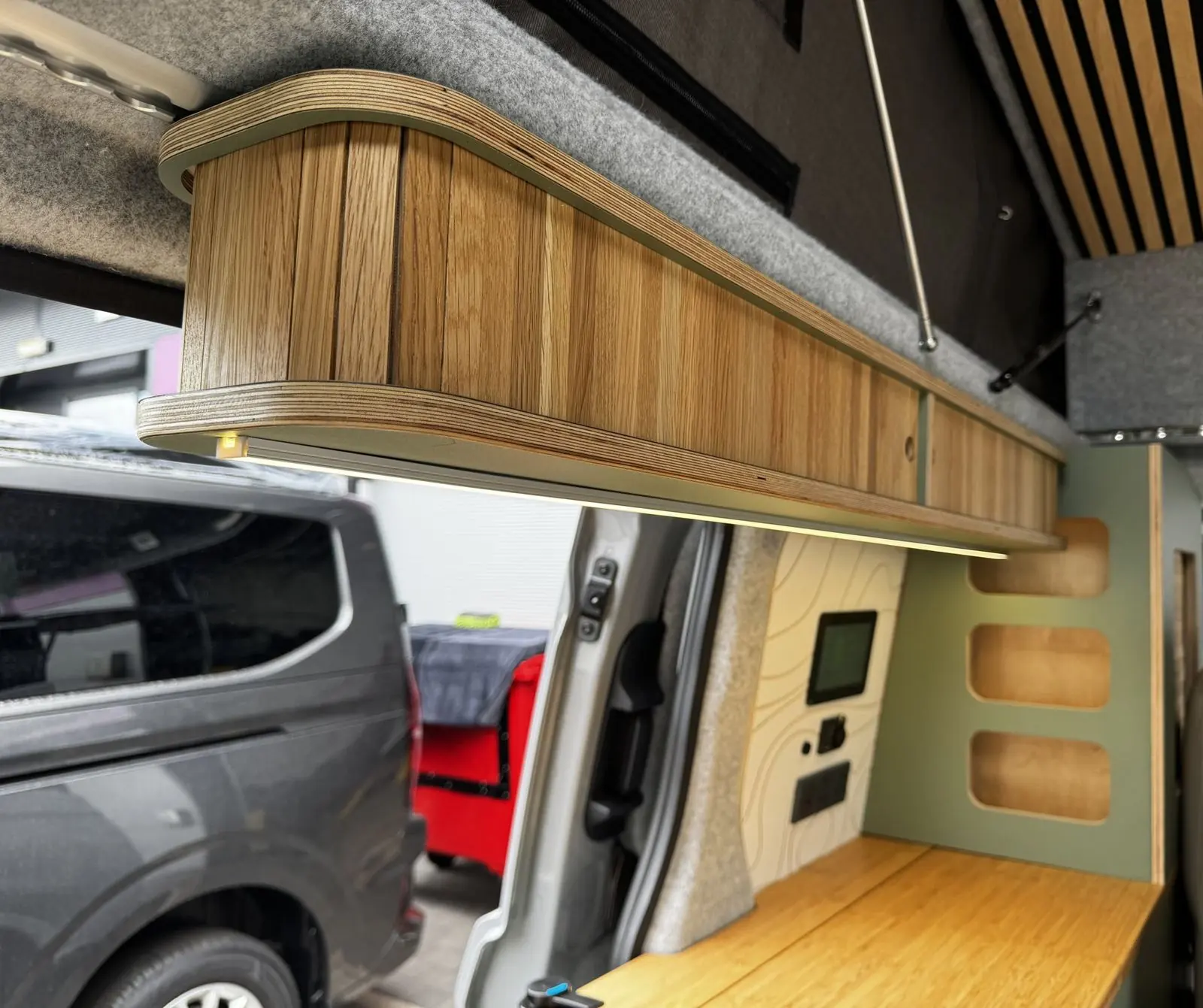 Oak overhead locker with rounded plywood ends and an LED strip underneath, with the slatted wood roof panel above and the sage green shelving and touchscreen panel below.