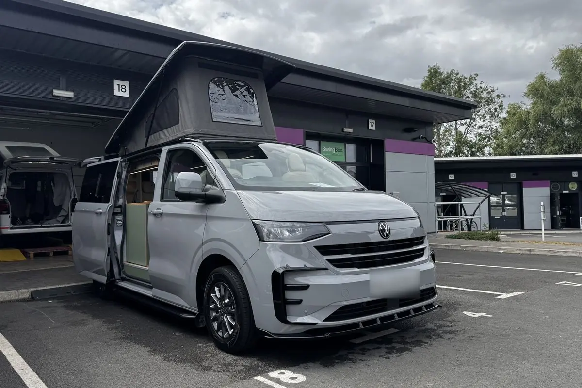 Front three-quarter view of a light grey new-shape Volkswagen Transporter campervan parked outside a workshop unit, with its dark grey pop-top roof raised, a side door slid open and a black front splitter.