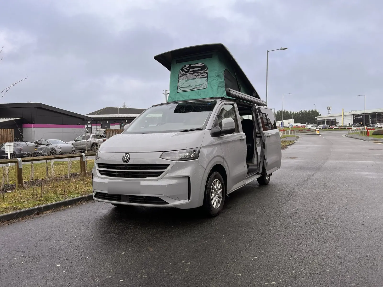 Front three-quarter view of a light grey new-shape Volkswagen Transporter campervan on a wet road with its pop-top roof raised, showing green canvas, and the side door open.