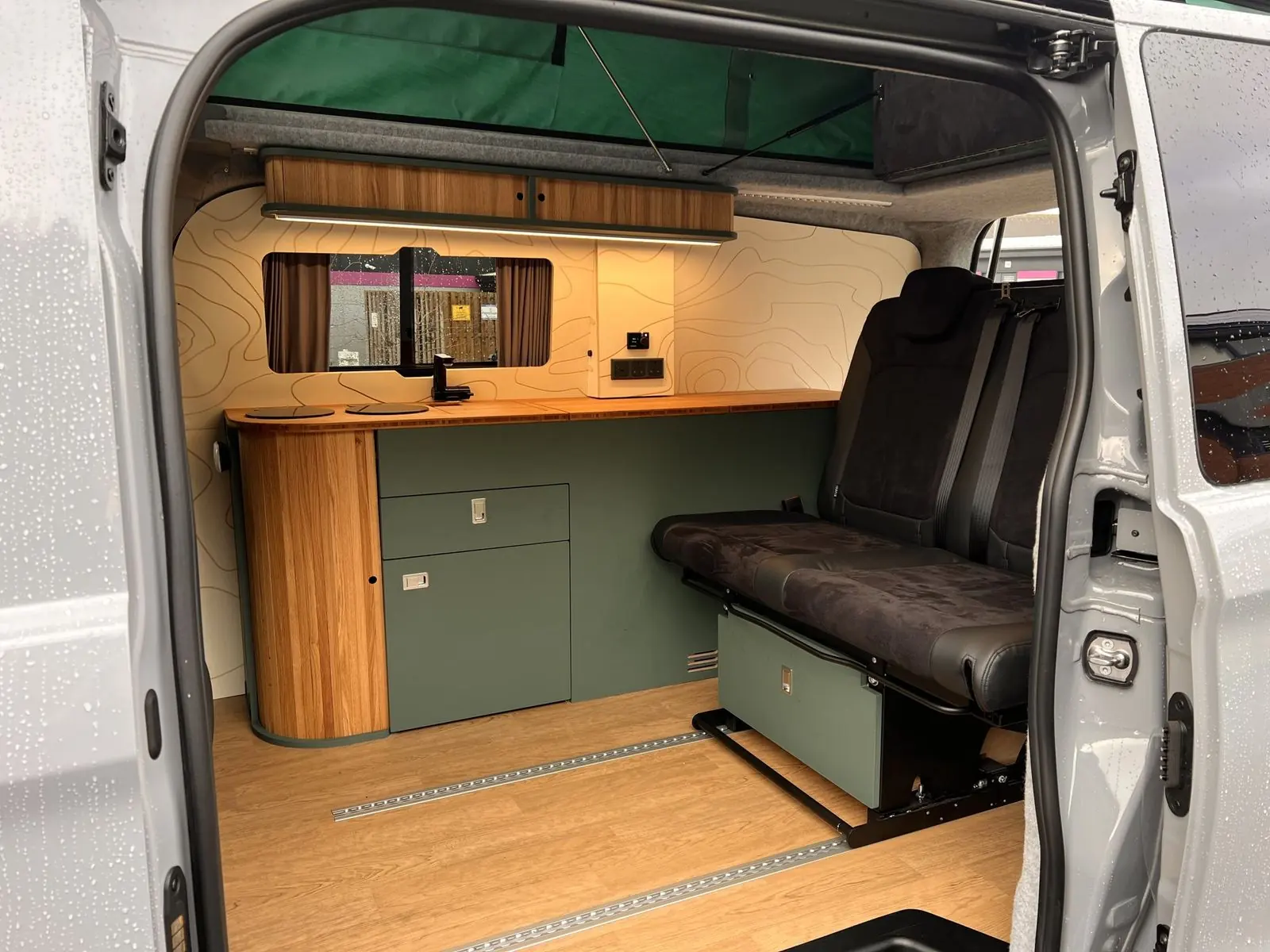 Interior seen through the side door: dark green kitchen unit with an oak slatted curved end and bamboo worktop, a two-seat rear bench in black suede-effect fabric, an oak overhead locker with LED strip, cream contour-line wall panels and green pop-top canvas above.