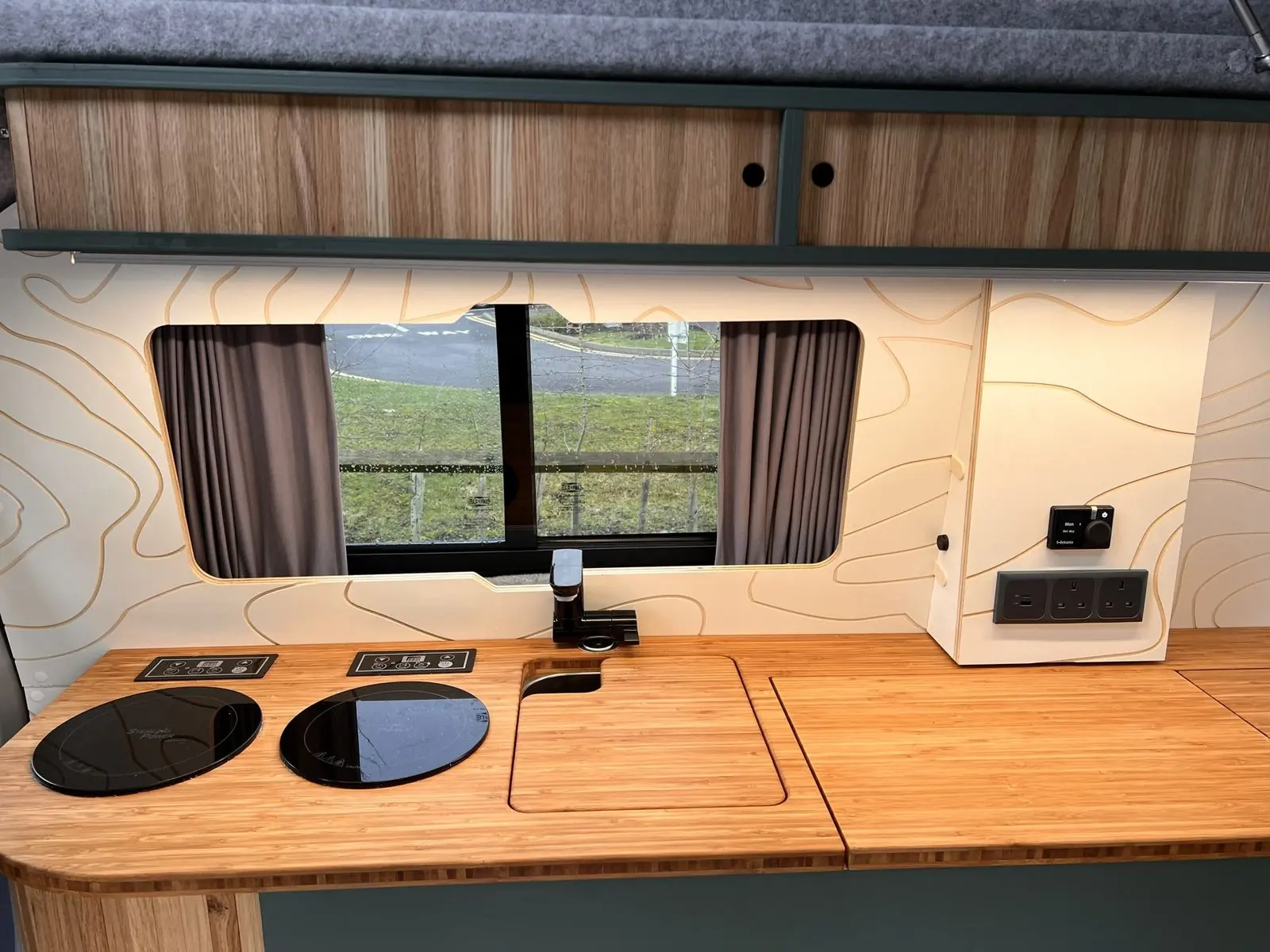 Kitchen from the front: bamboo worktop with two round black induction hobs, a flush bamboo sink cover and black tap, a window with grey curtains, an oak overhead locker and a wall panel with a dial controller and sockets.
